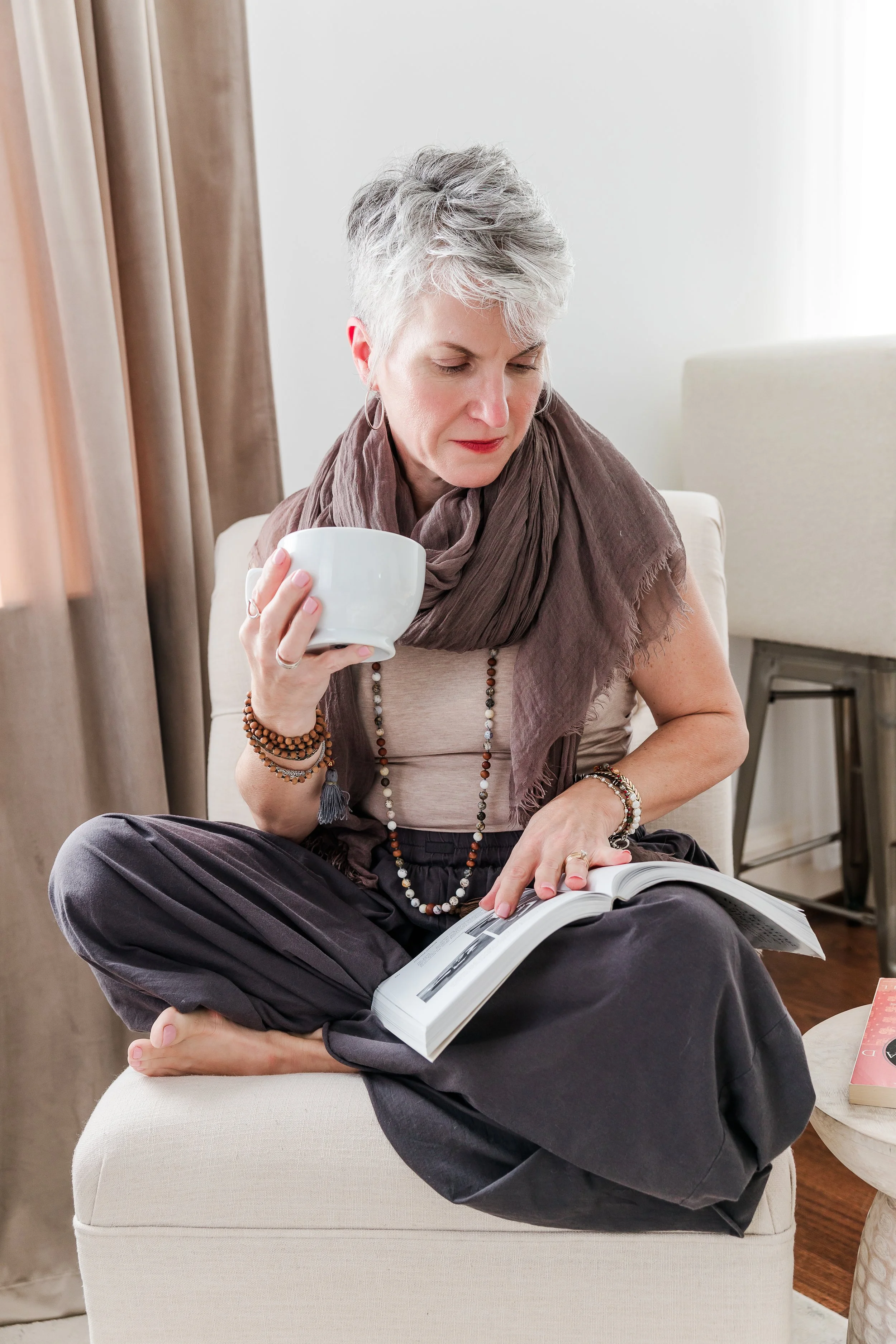 Heather Hatcher with short gray hair sitting cross-legged on an ottoman, holding a mug while reading in a North Carolina studio.