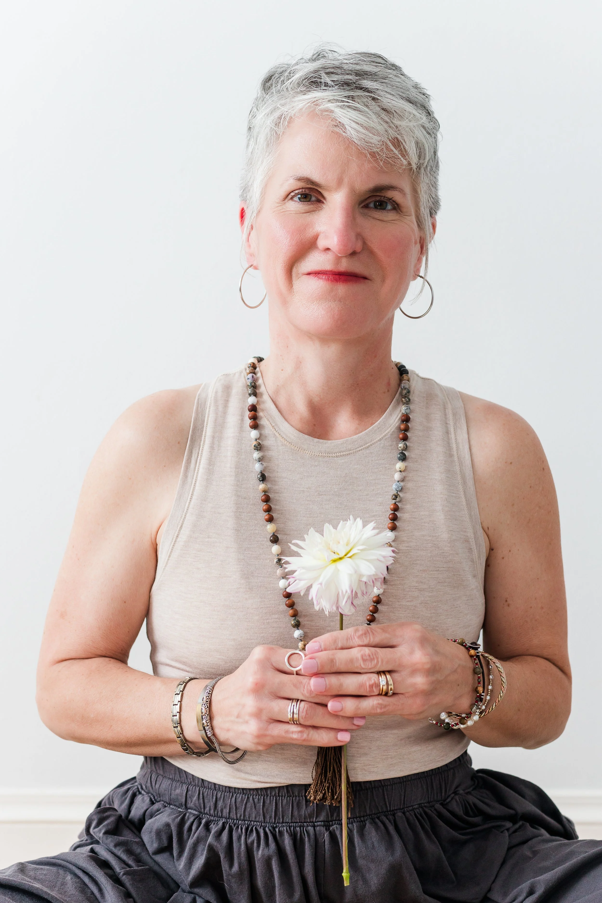 Heather Hatcher with short gray hair wearing a beige sleeveless top and black skirt, holding a white flower in a North Carolina studio setting.