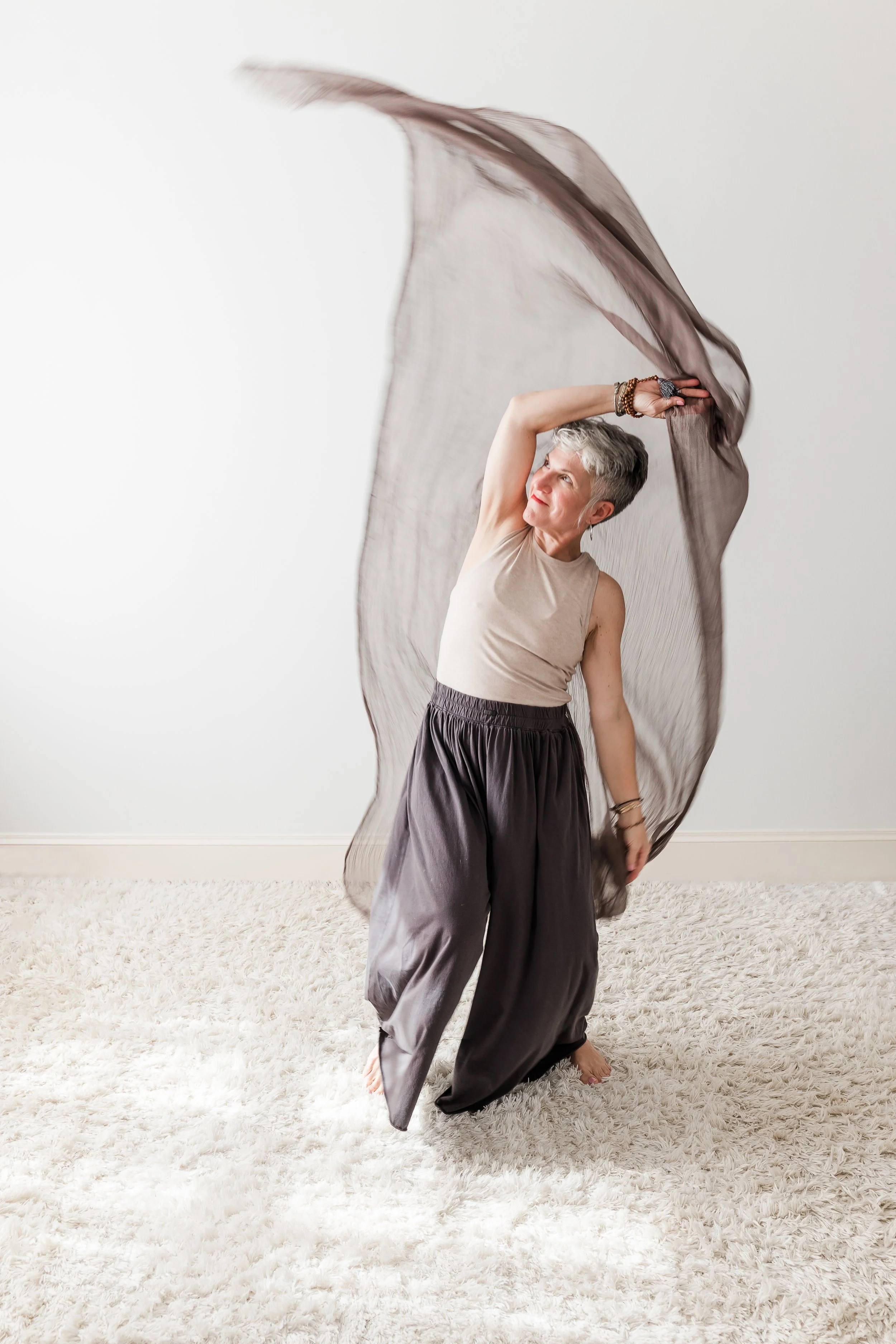 Heather Hatcher dancing with a sheer brown scarf, wearing a beige sleeveless top and dark pants in a minimal white space.