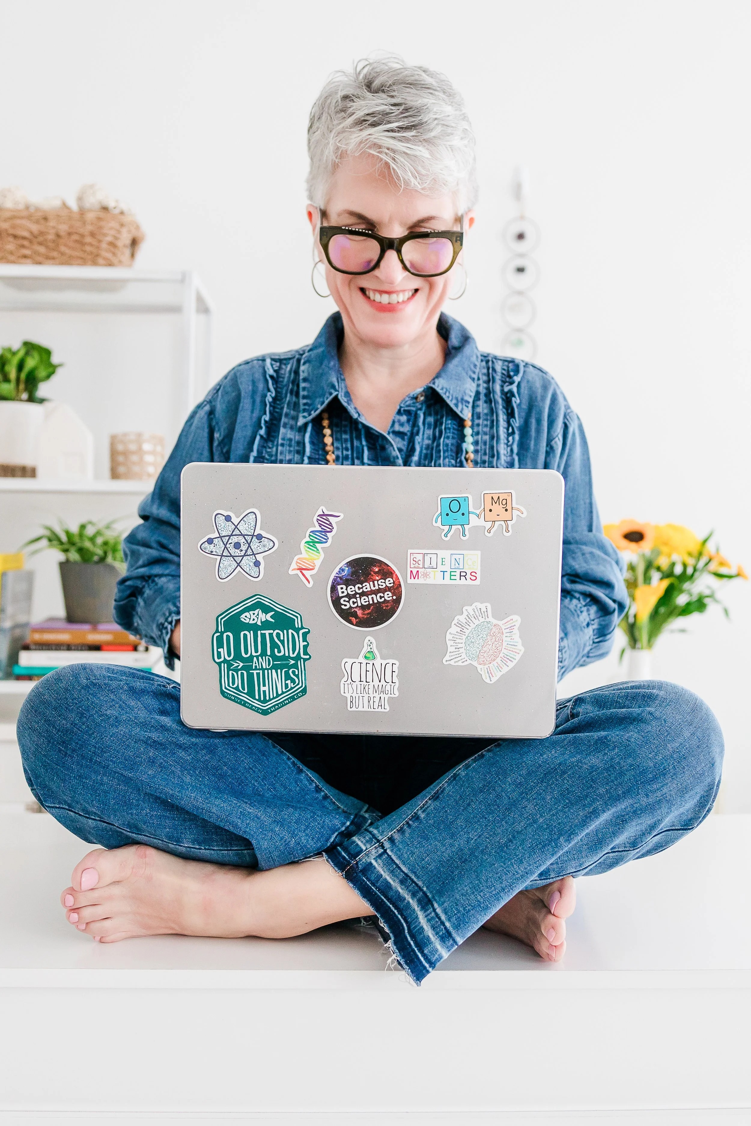 Heather Hatcher with short gray hair and glasses, sitting cross-legged with a laptop in a bright North Carolina studio space.