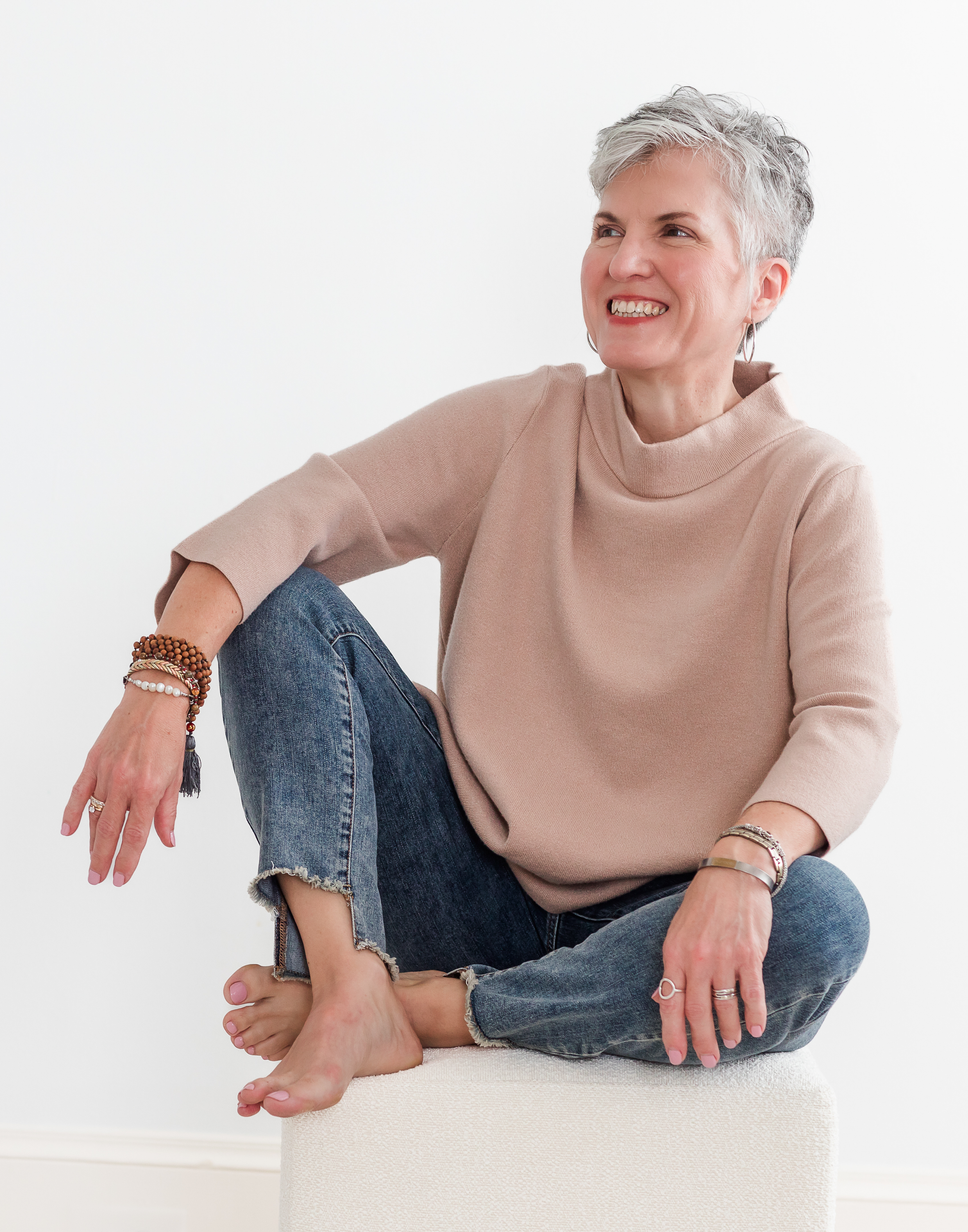 Heather Hatcher with short gray hair smiling while seated cross-legged on a white ottoman in a North Carolina studio setting.