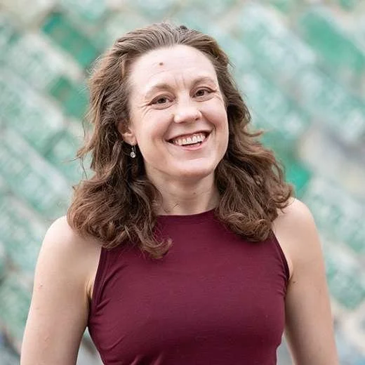 A smiling woman with wavy brown hair wearing a sleeveless maroon top against a blurred outdoor background.