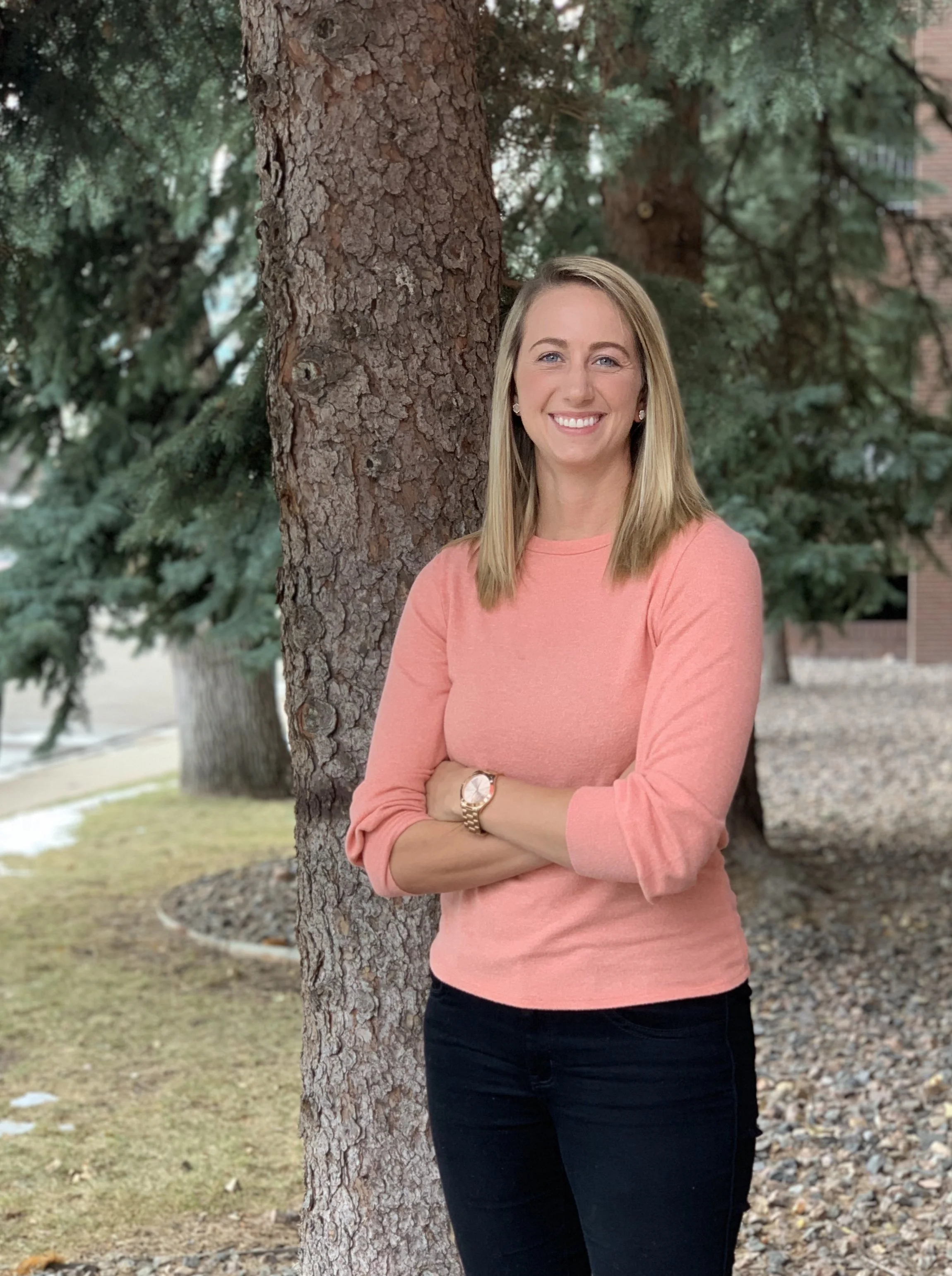 A woman with shoulder length blonde hair, wearing a pink sweater, black pants, and a watch, smiling and standing outdoors with her arms crossed against a tree.