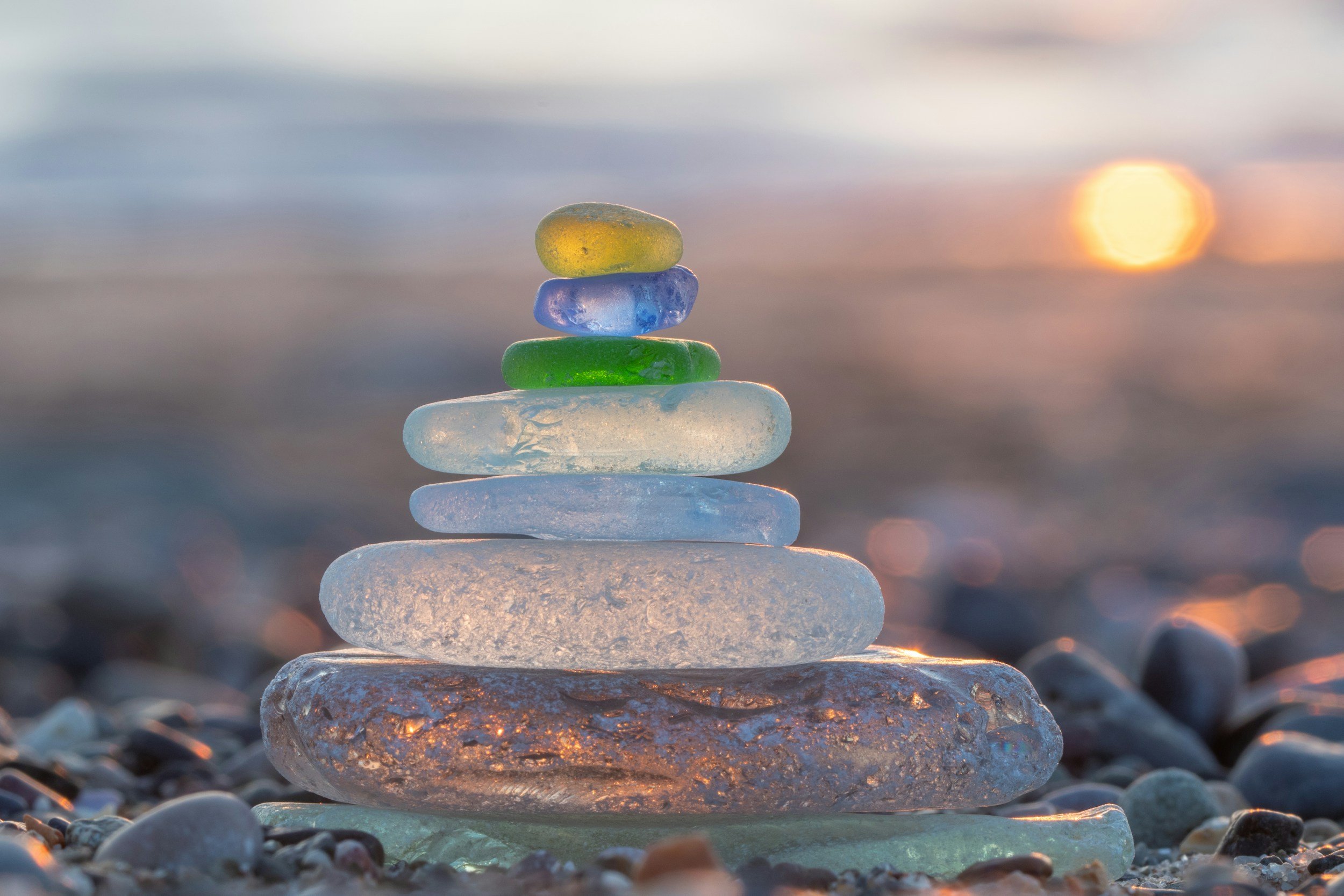 Colorful glass stones stacked in a pyramid shape on a rocky beach during sunset or sunrise.