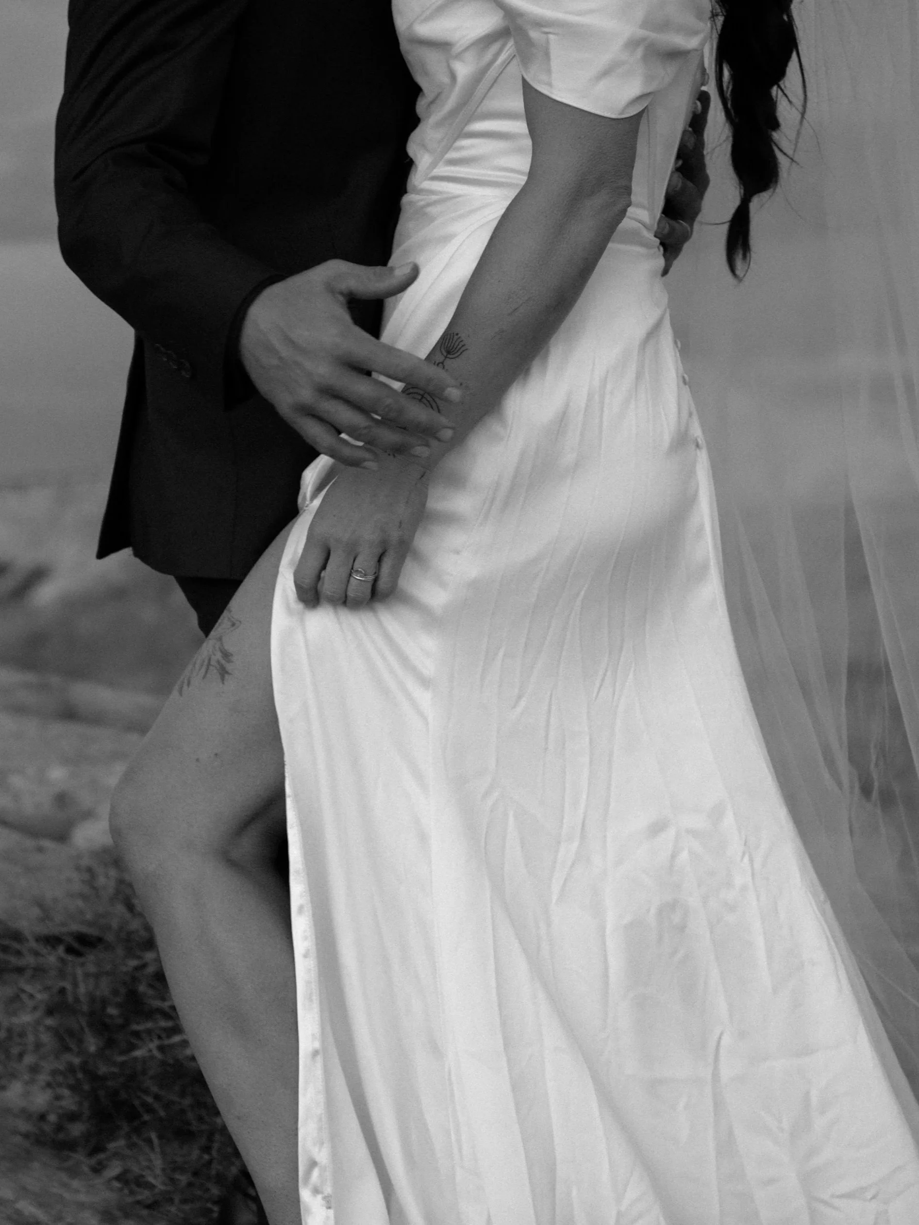 A close-up black and white photo of a couple on their wedding day, with the man touching the woman's waist and hand, highlighting wedding rings and tattoos on her arm and thigh.