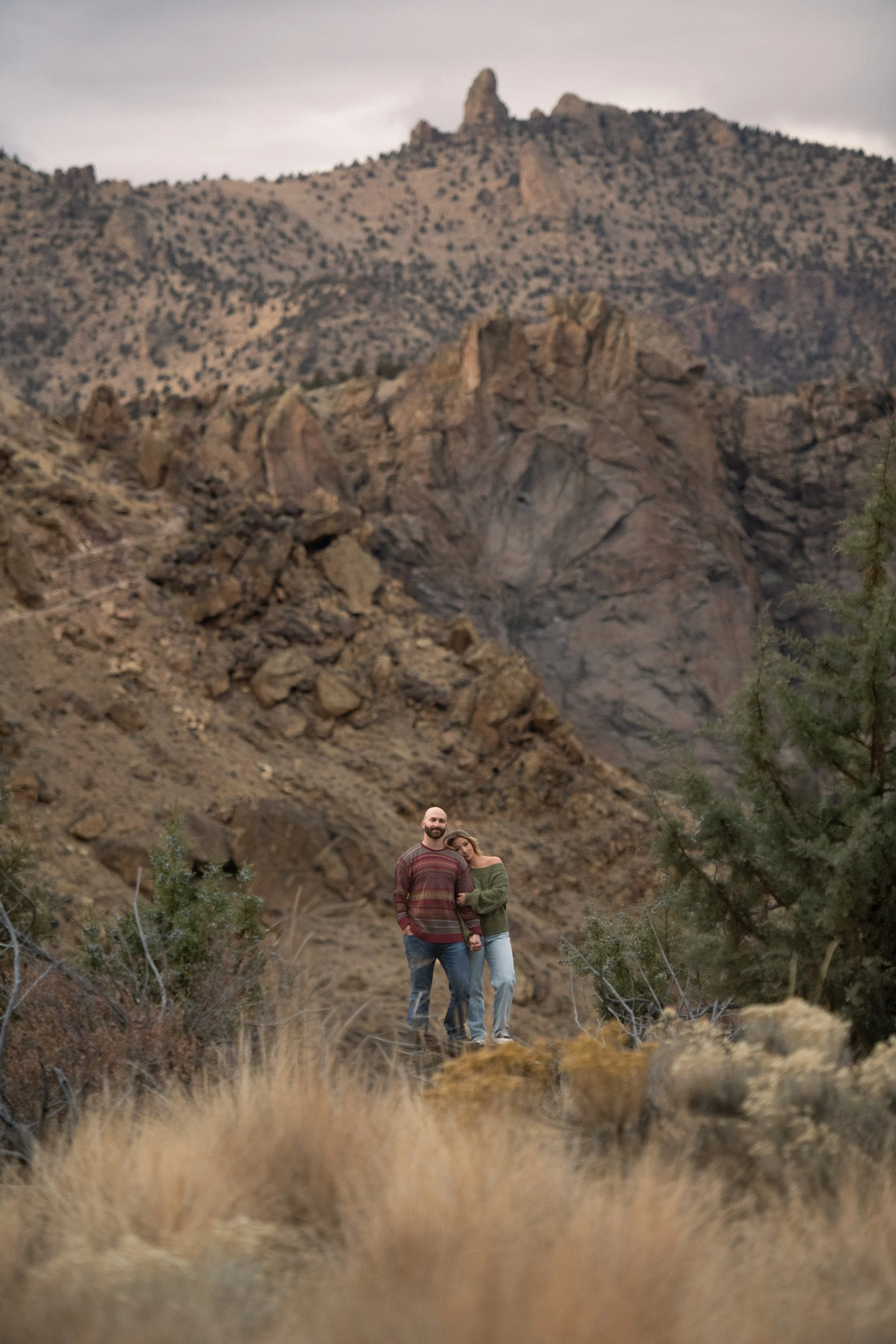 A couple standing hand-in-hand in a rocky desert landscape with mountains and sparse vegetation in the background in Smith Rock State Park.