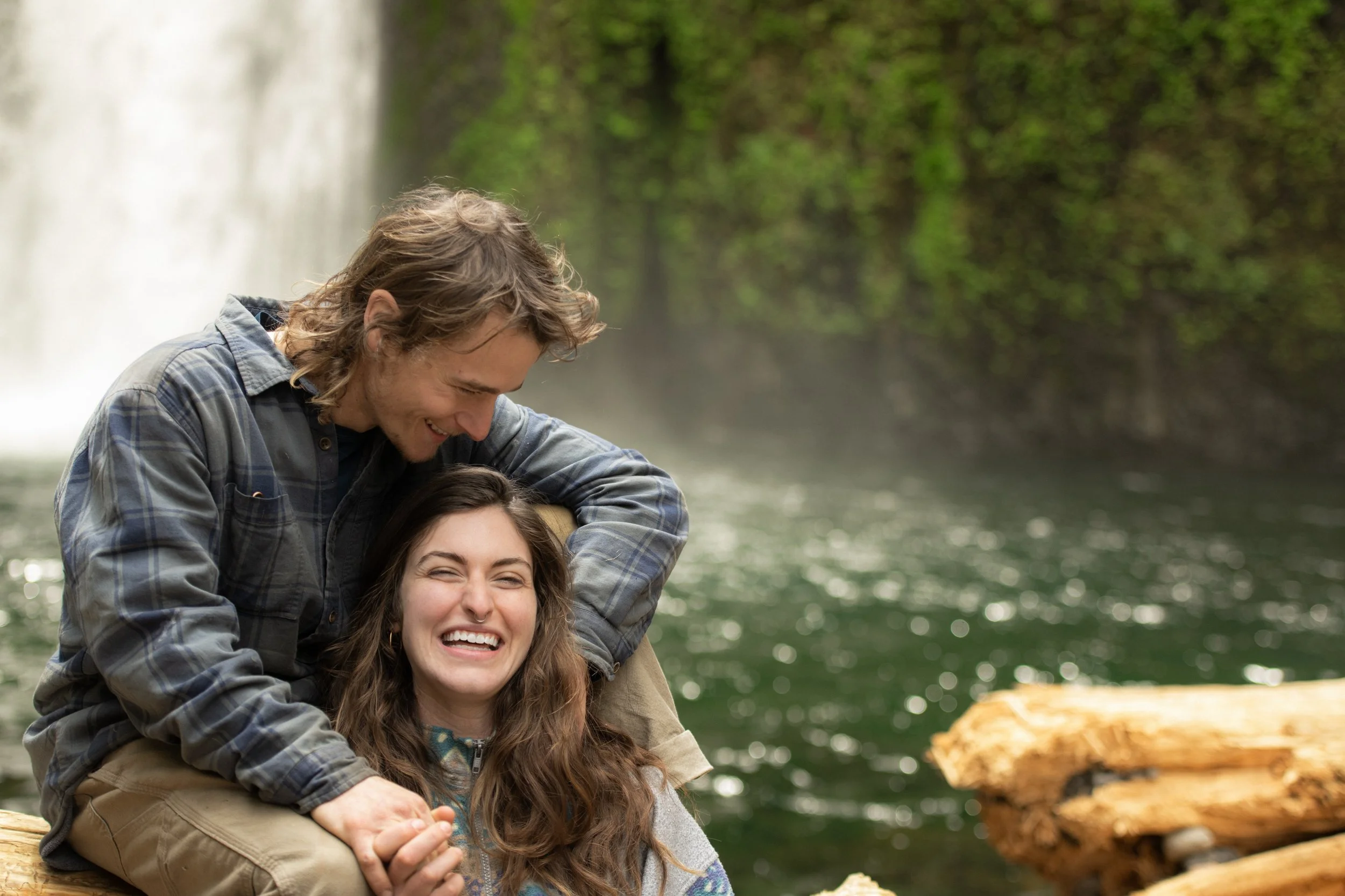 Two people smiling and laughing near a river in a forested area with a waterfall in the background in the Columbia River Gorge