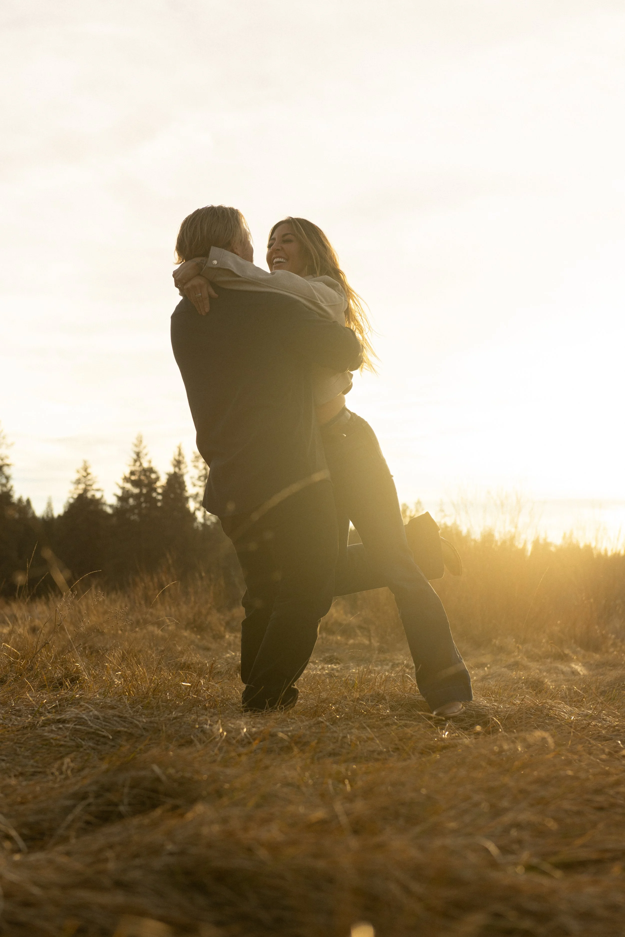 A couple hugging outdoors during sunset, with one lifting the other, in a grassy field with trees in the background in Central Oregon.