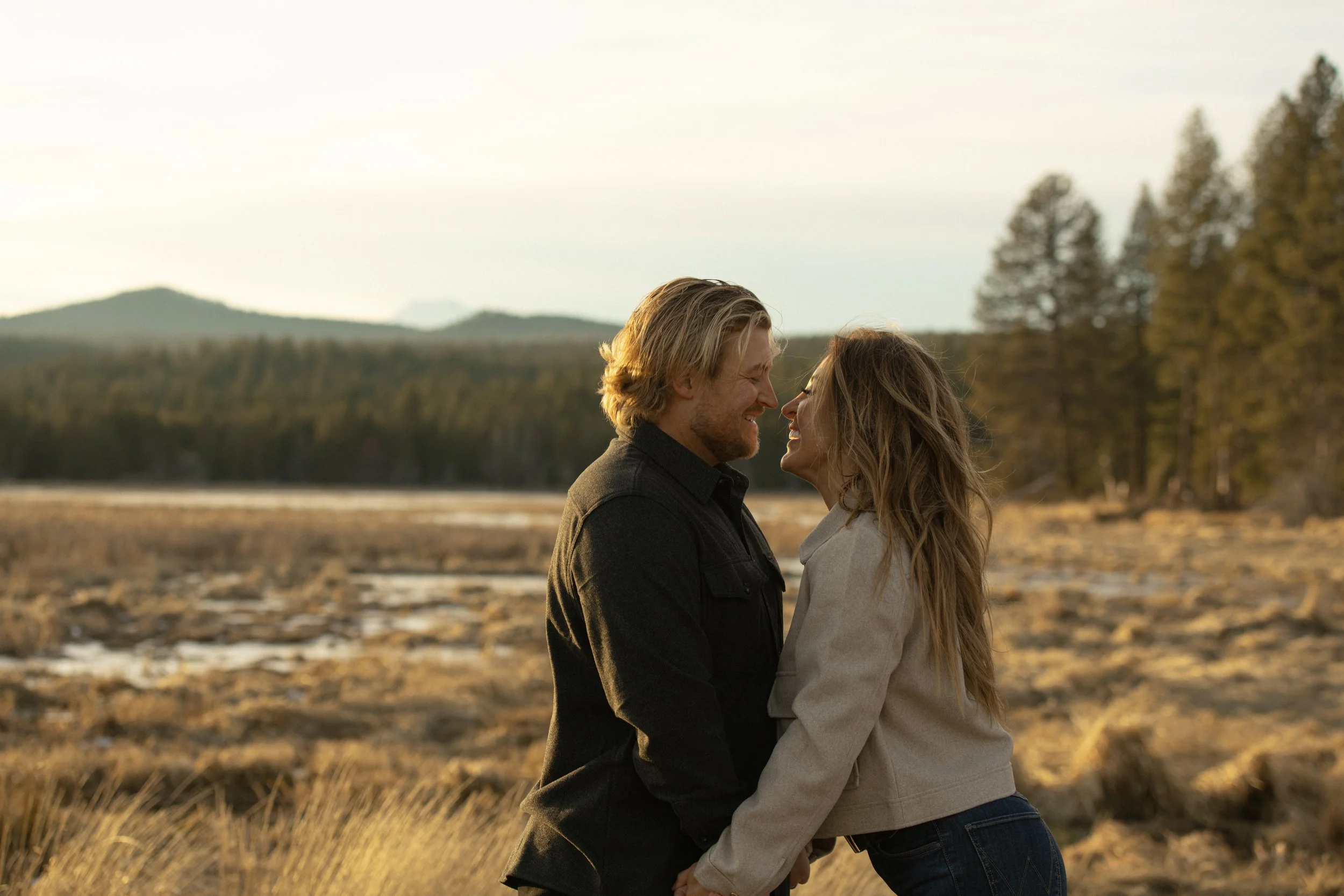 A couple stands in a natural outdoor setting, smiling and leaning close to each other amid a field with trees and hills in the background during sunset in Bend, Oregon.