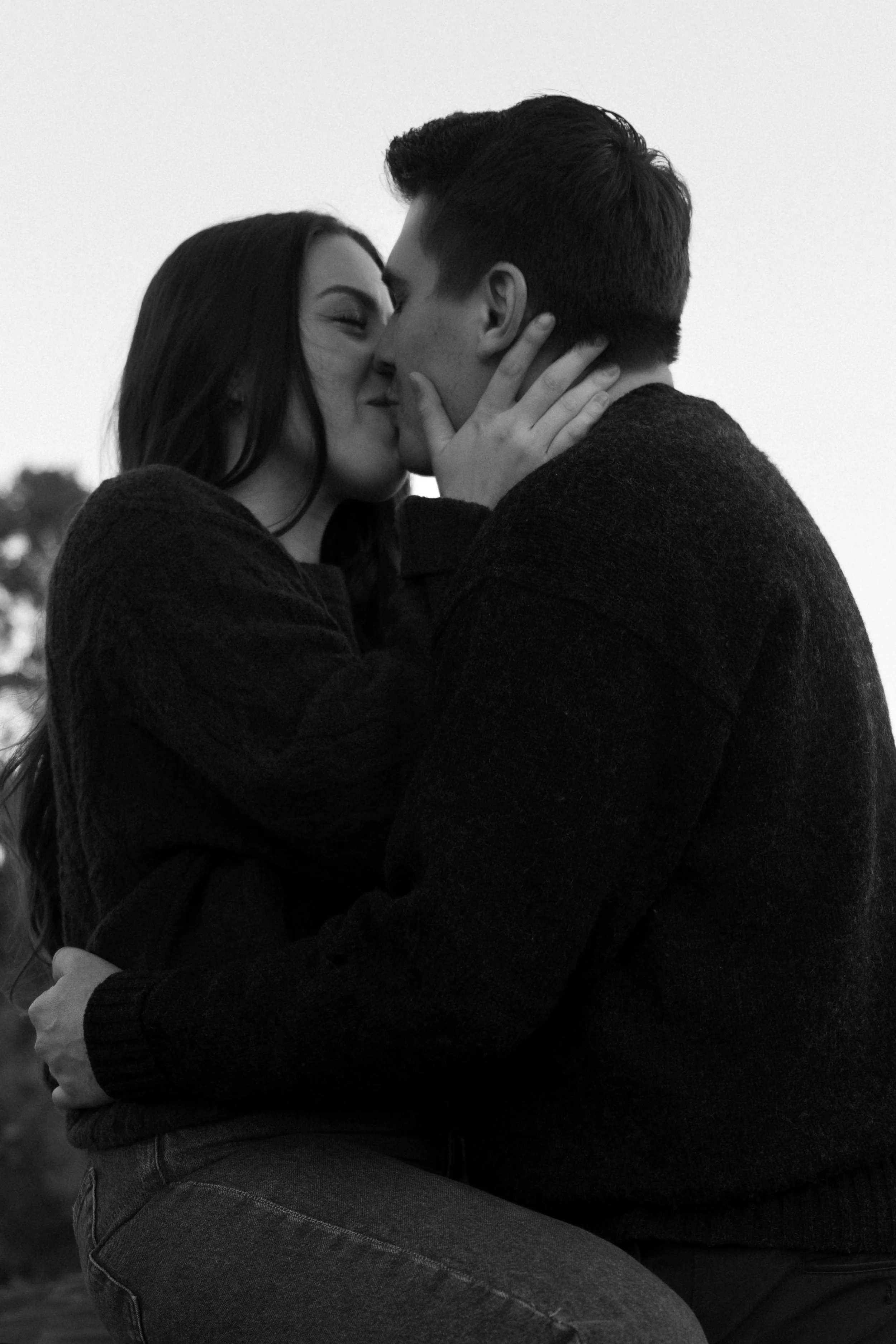 A black-and-white photo of a couple kissing, with the woman's hand touching the man's face. The woman has long dark hair, and the man has short dark hair. They are dressed in dark sweaters, and the background is blurred with trees visible.