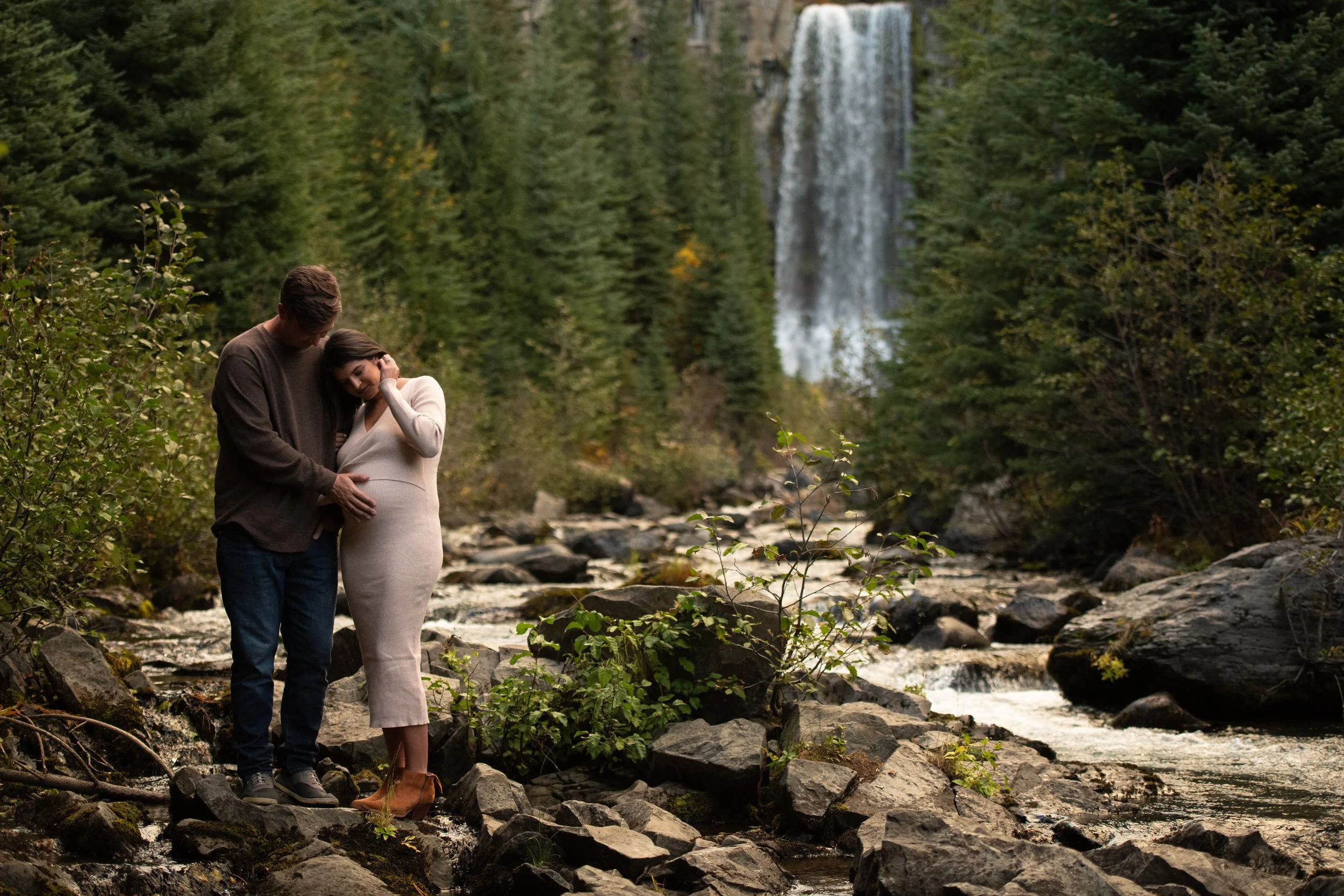 A pregnant woman and a man standing together on rocks near a stream with Tumalo waterfall in the background, surrounded by green trees.