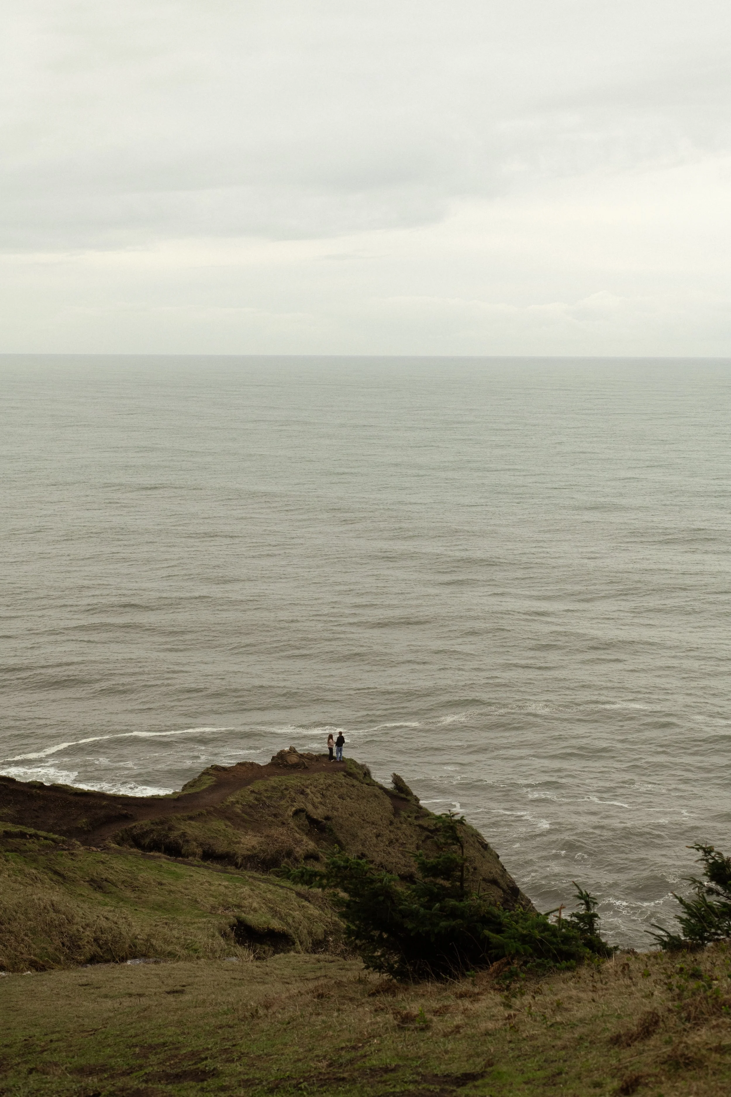 Two people standing on a grassy cliff overlooking the ocean under a cloudy sky on the Oregon Coast.
