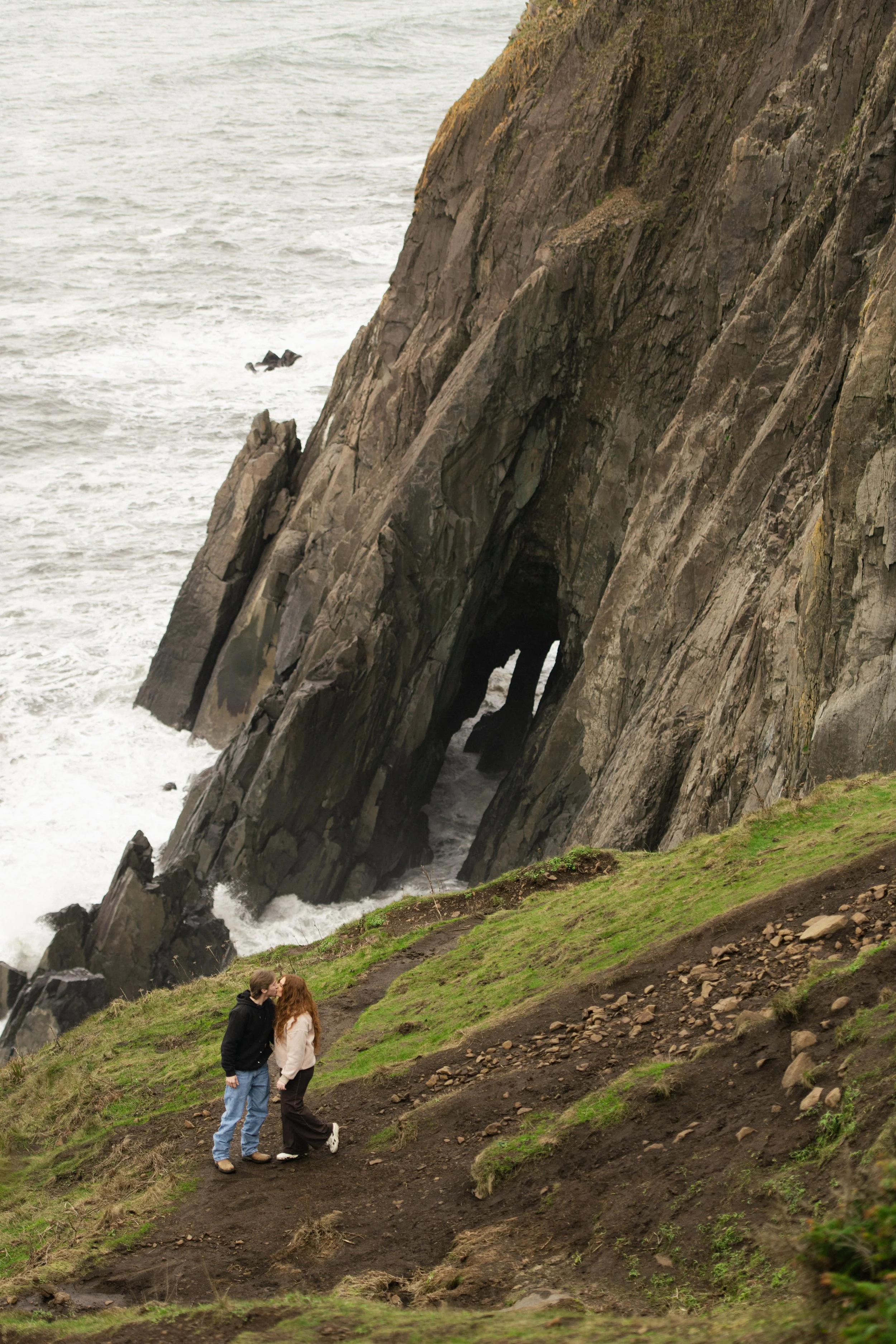 A couple standing on a grassy dirt trail near the rocky Oregon coastline with a large arch-shaped rock formation and ocean waves in the background.