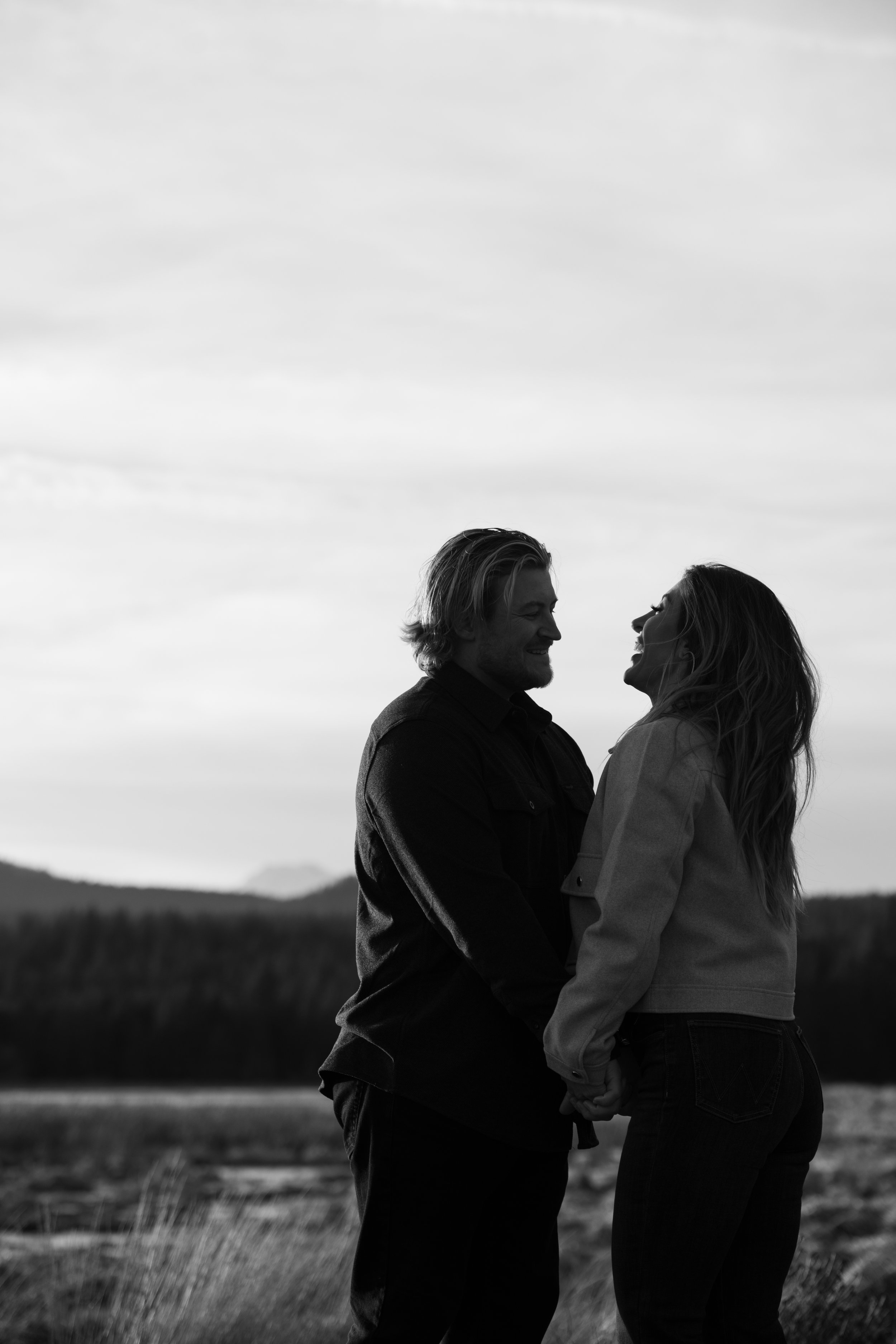 A black and white photo of a couple holding hands and smiling at each other outdoors with a mountainous landscape and cloudy sky in the background.