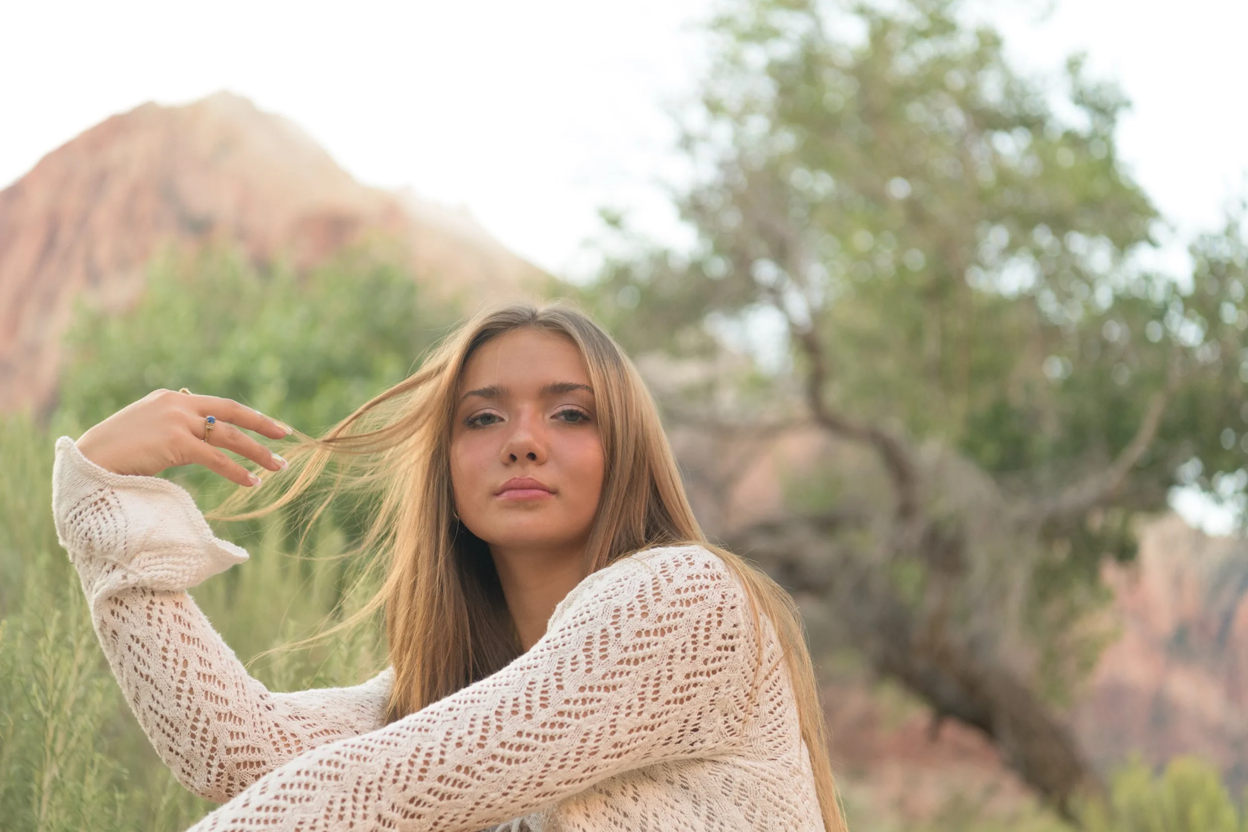 A young woman with long, light brown hair and a neutral expression, wearing a cream-colored knit sweater, outdoors with greenery and Zion National Park in the background.