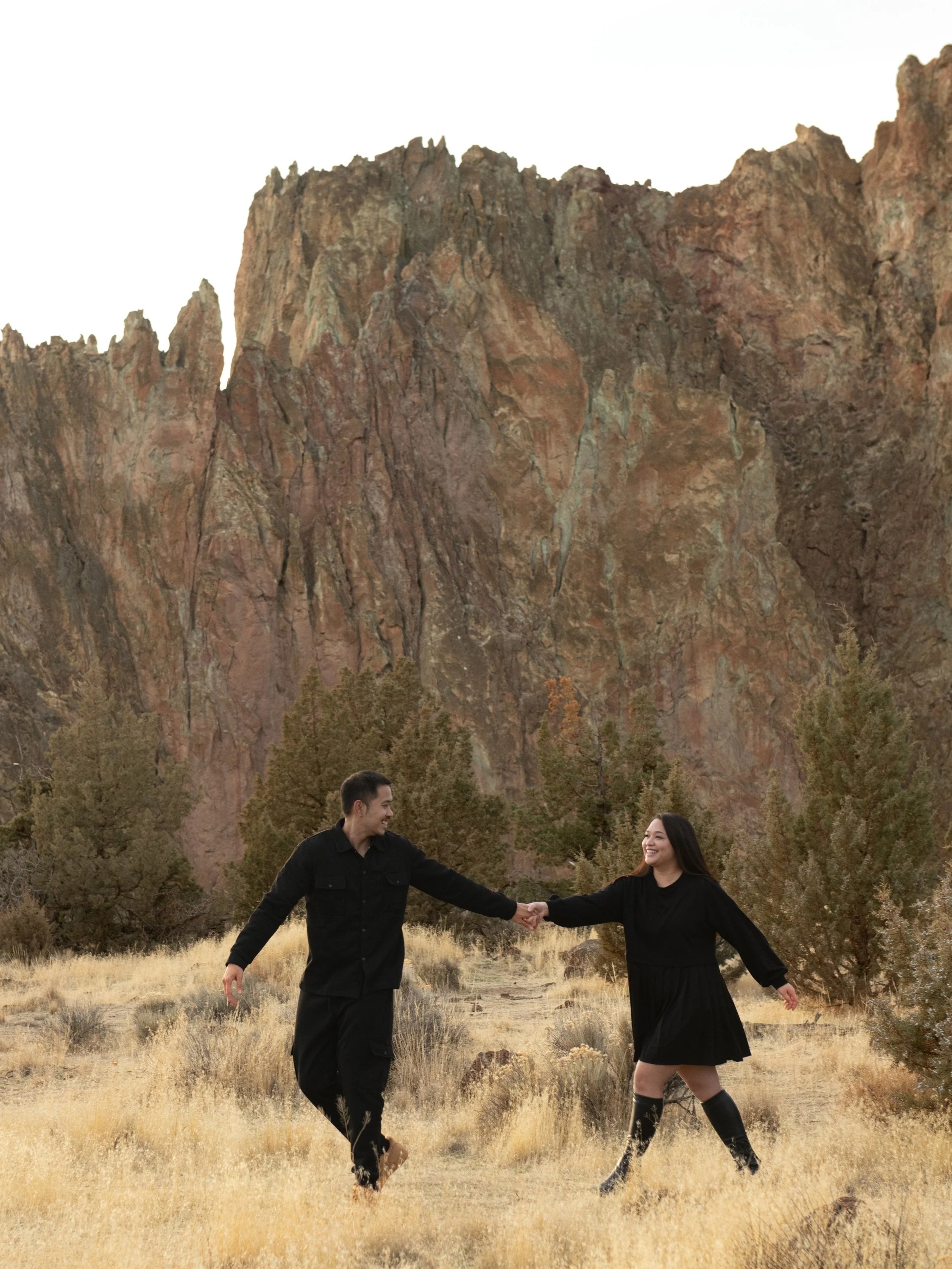 A man and woman holding hands, walking through a dry grassy field with trees and large rocky cliffs in the background, smiling at each other at Smith Rock State Park.