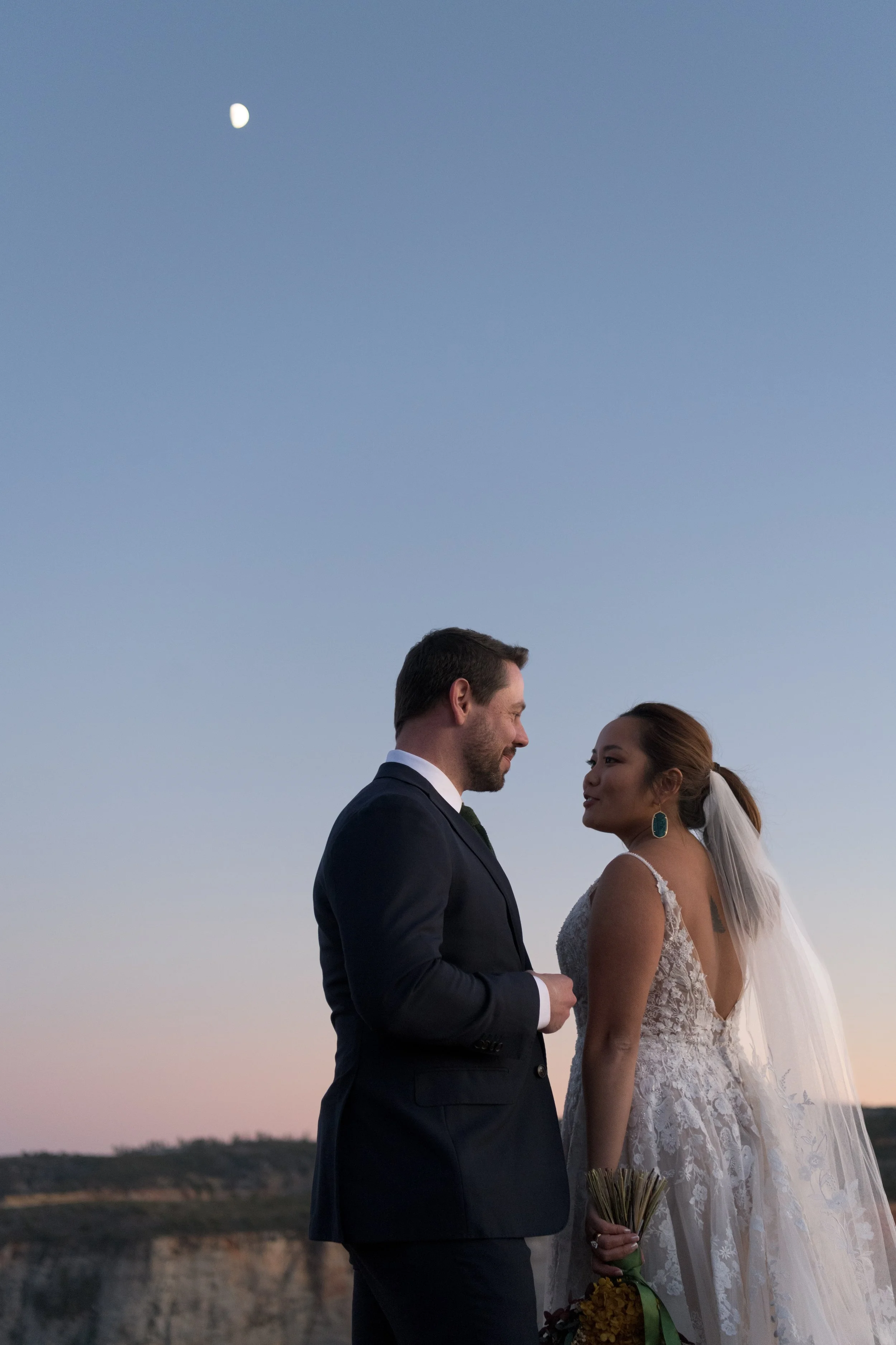 A bride and groom standing close together outdoors during a wedding at sunset or dusk, with the moon visible in the sky.