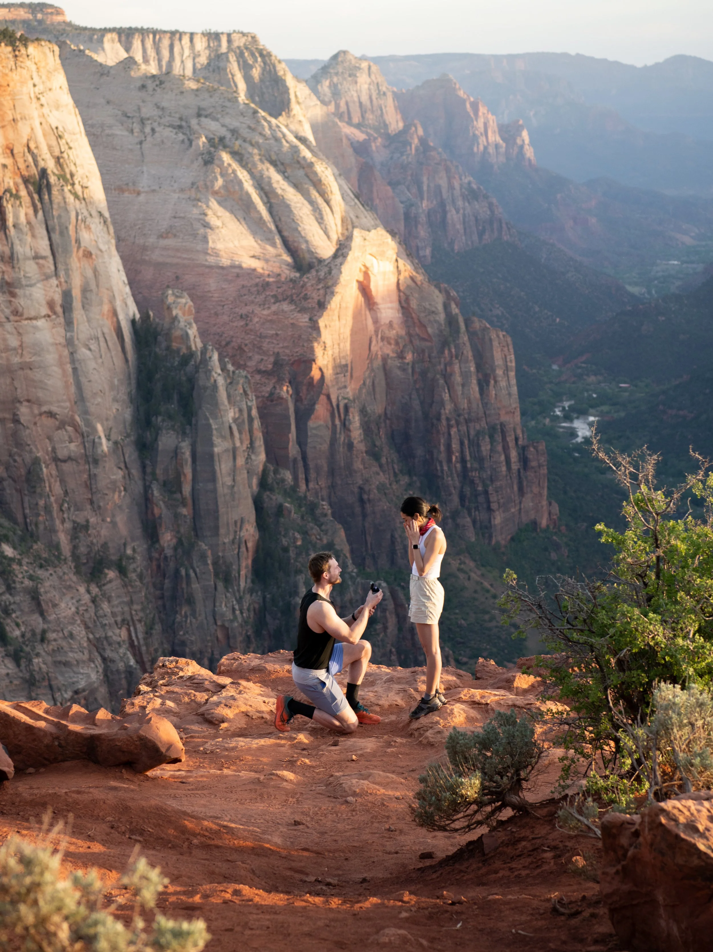 A man proposing marriage to a woman on a rocky cliff in Zion National Park with towering red and gray cliffs in the background, during sunset.