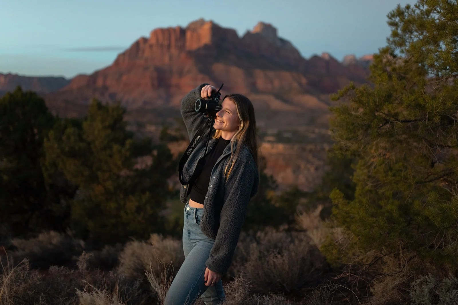 A woman in a black crop top, gray jacket, and jeans standing outdoors near juniper trees with Zion National Park in the background, smiling and holding a camera to her face.