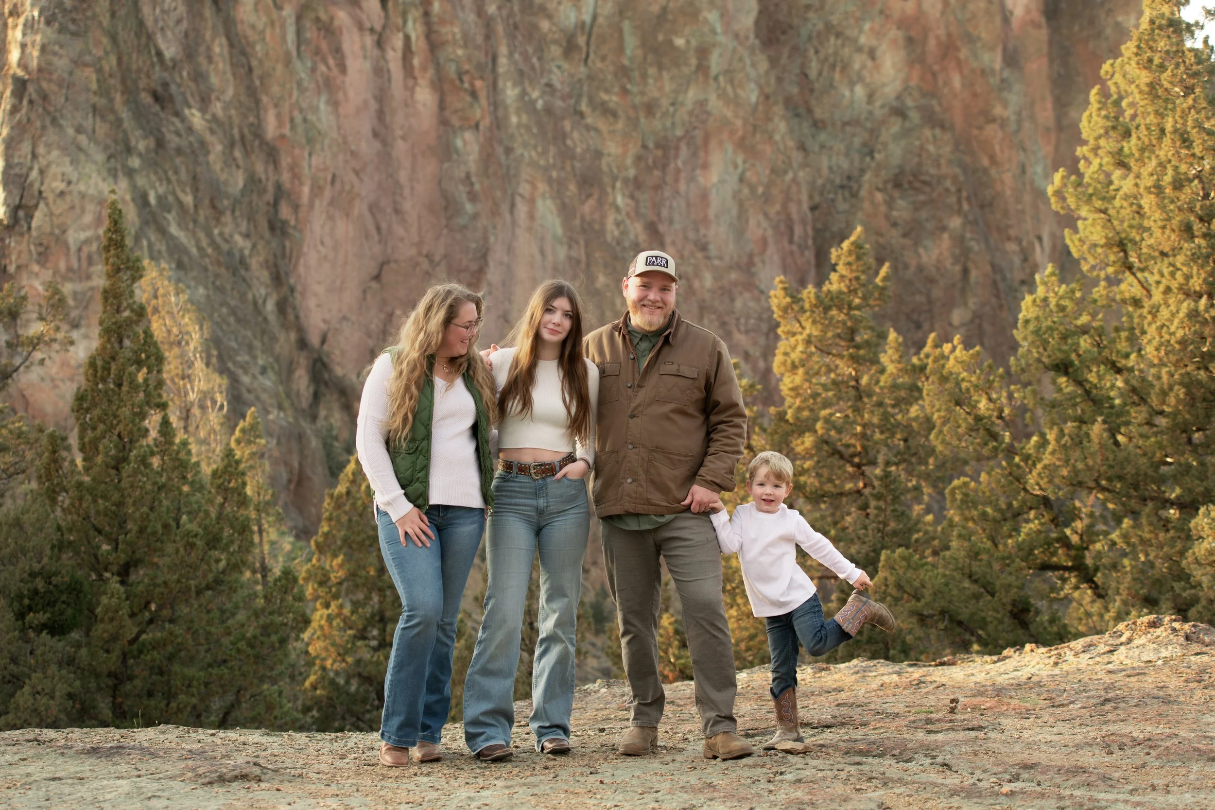 A family of five standing outdoors near trees and rocky cliffs, smiling at the camera in Smith Rock State Park.
