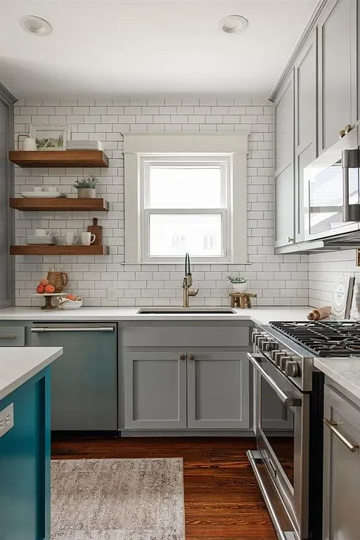 A modern kitchen with white subway tile backsplash, a window above the sink, light gray cabinets, wooden floating shelves with dishes and decor, and a stainless steel stove.