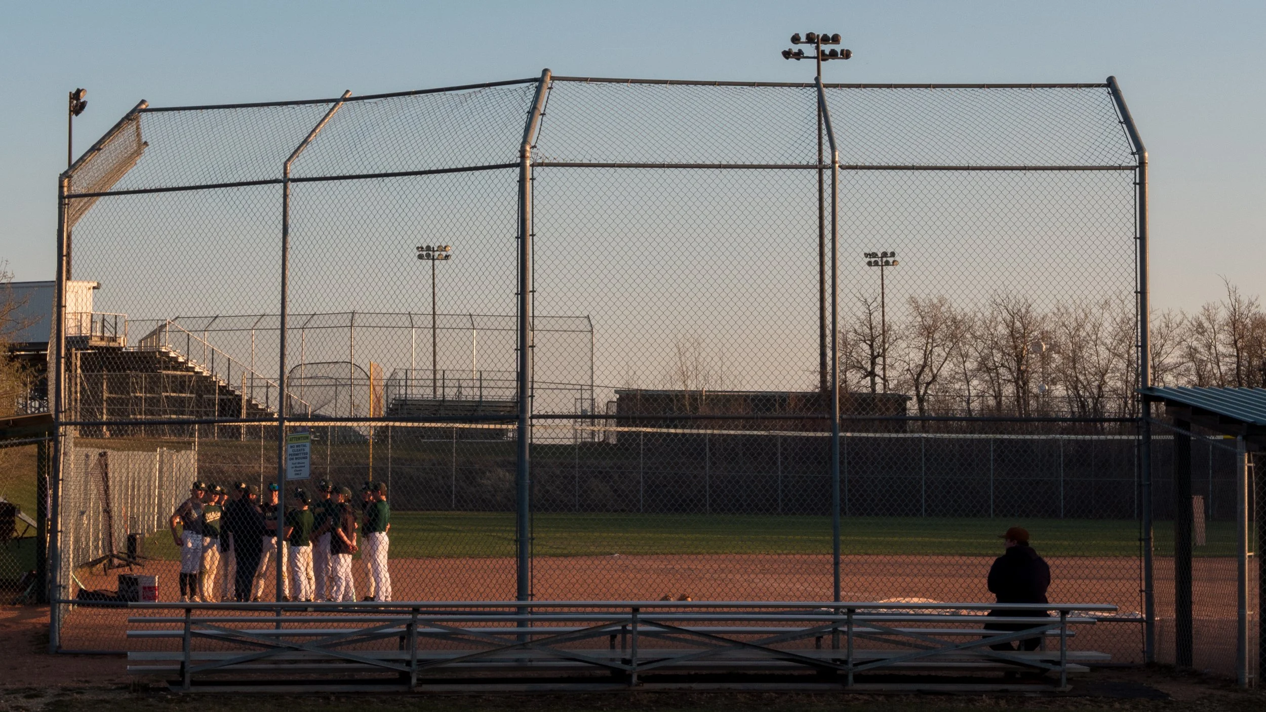 The image depicts a baseball field with a group of players and spectators, enclosed by a tall chain-link fence. The background features tall light poles and trees, under a clear sky during dusk.
