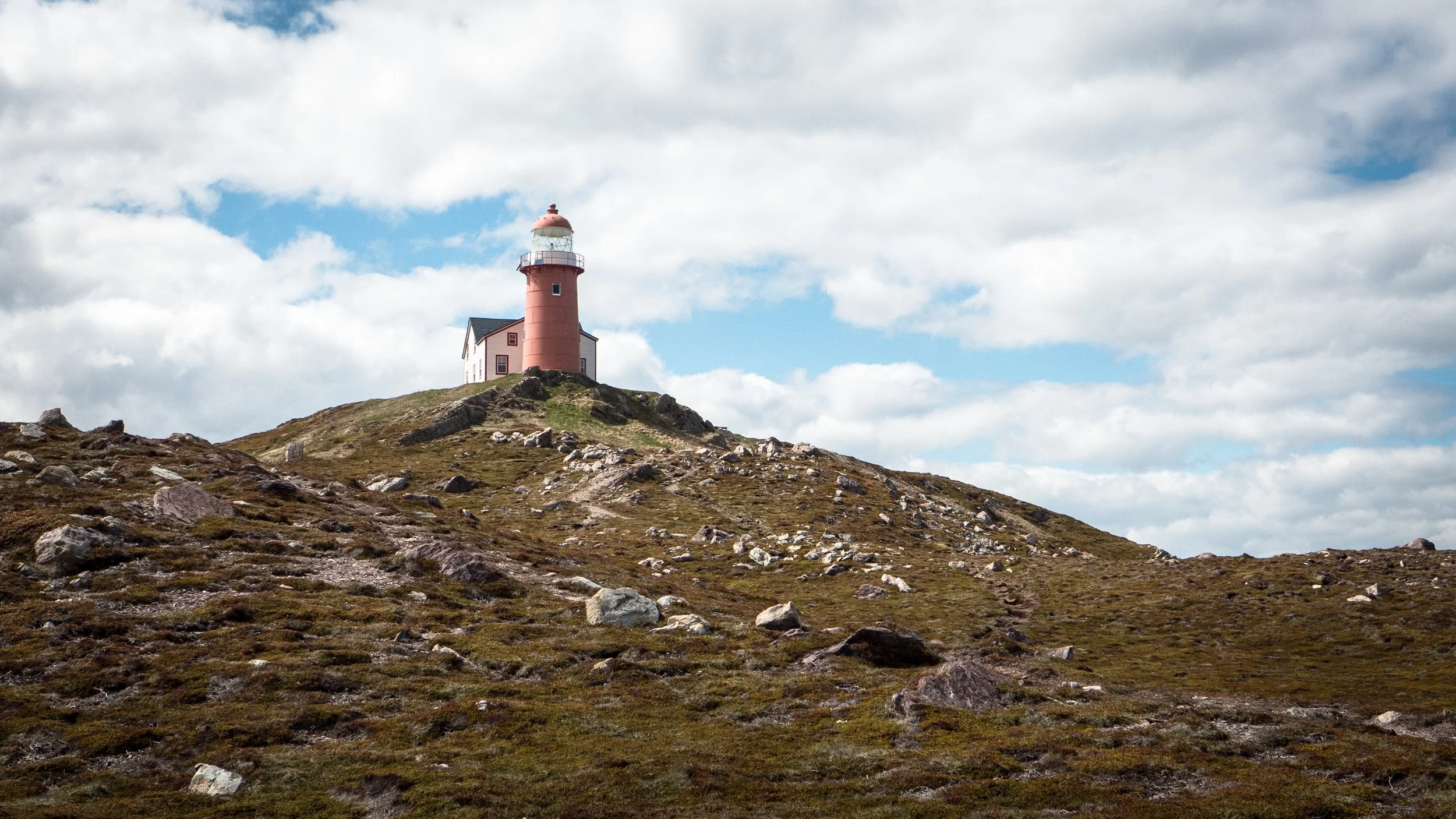 A lighthouse painted pink, situated on a grassy hill with rocks, under a partly cloudy sky.