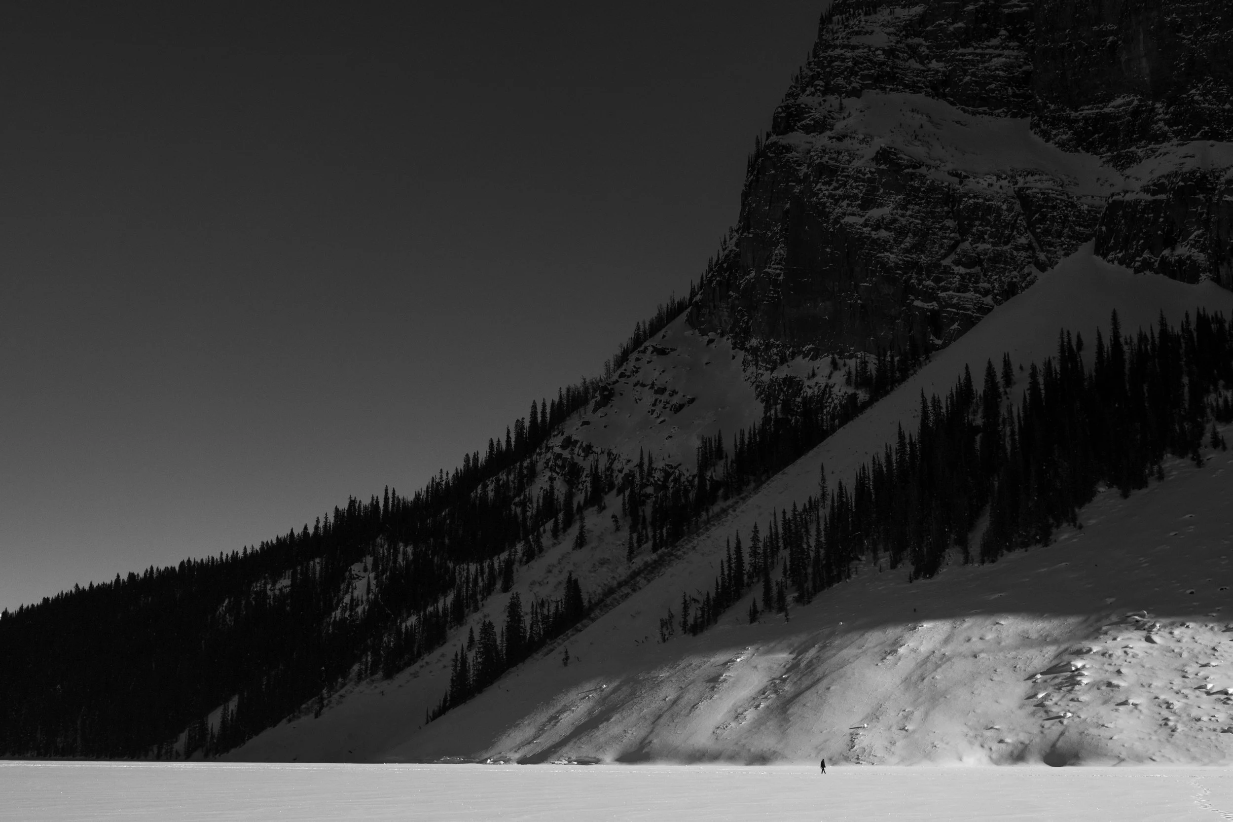A lone person standing on a vast snow-covered landscape, with a large mountain with snow and pine trees in the background under a clear sky.