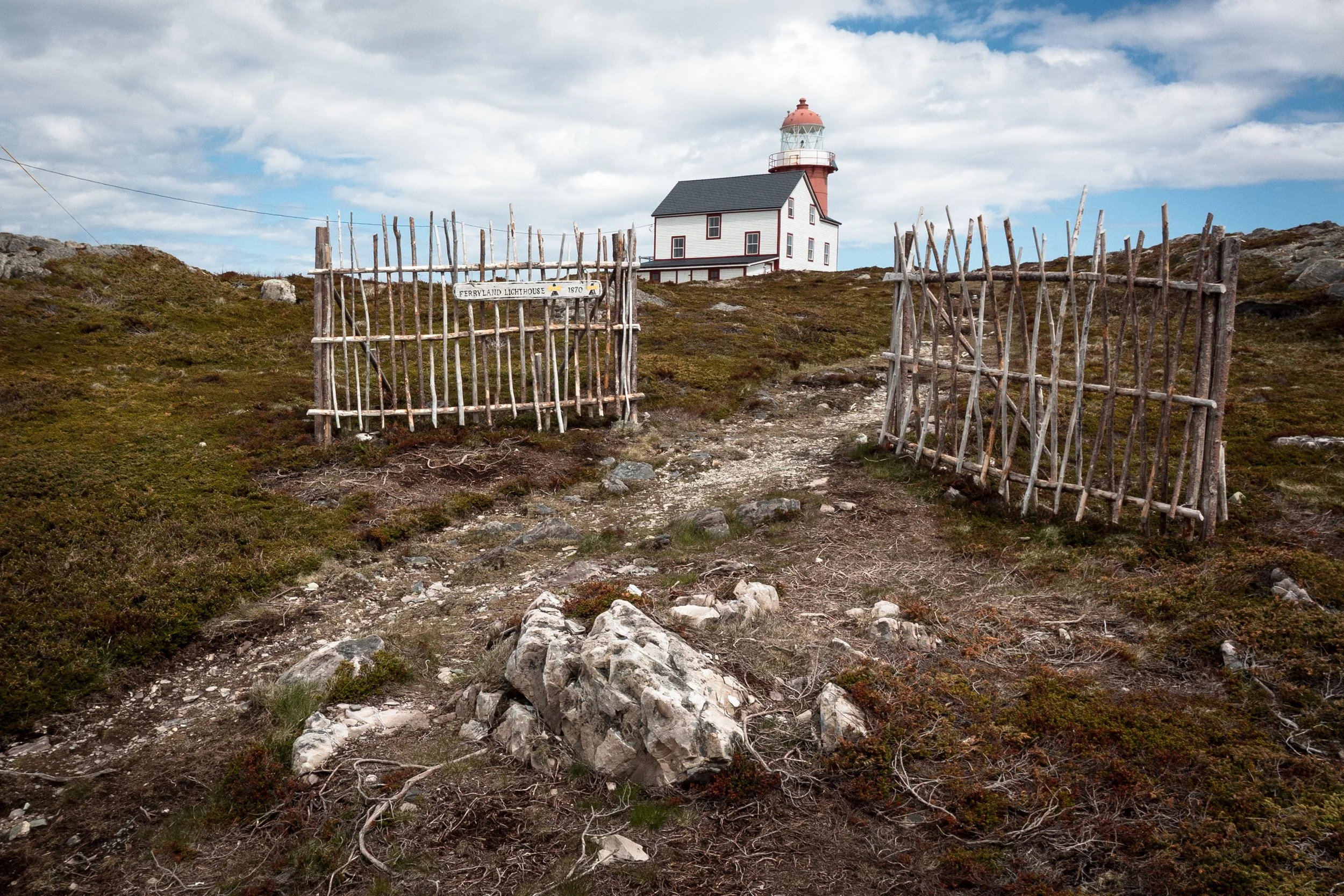 A lighthouse on a hill with a white building, enclosed by a rustic wooden fence, under a partly cloudy sky.