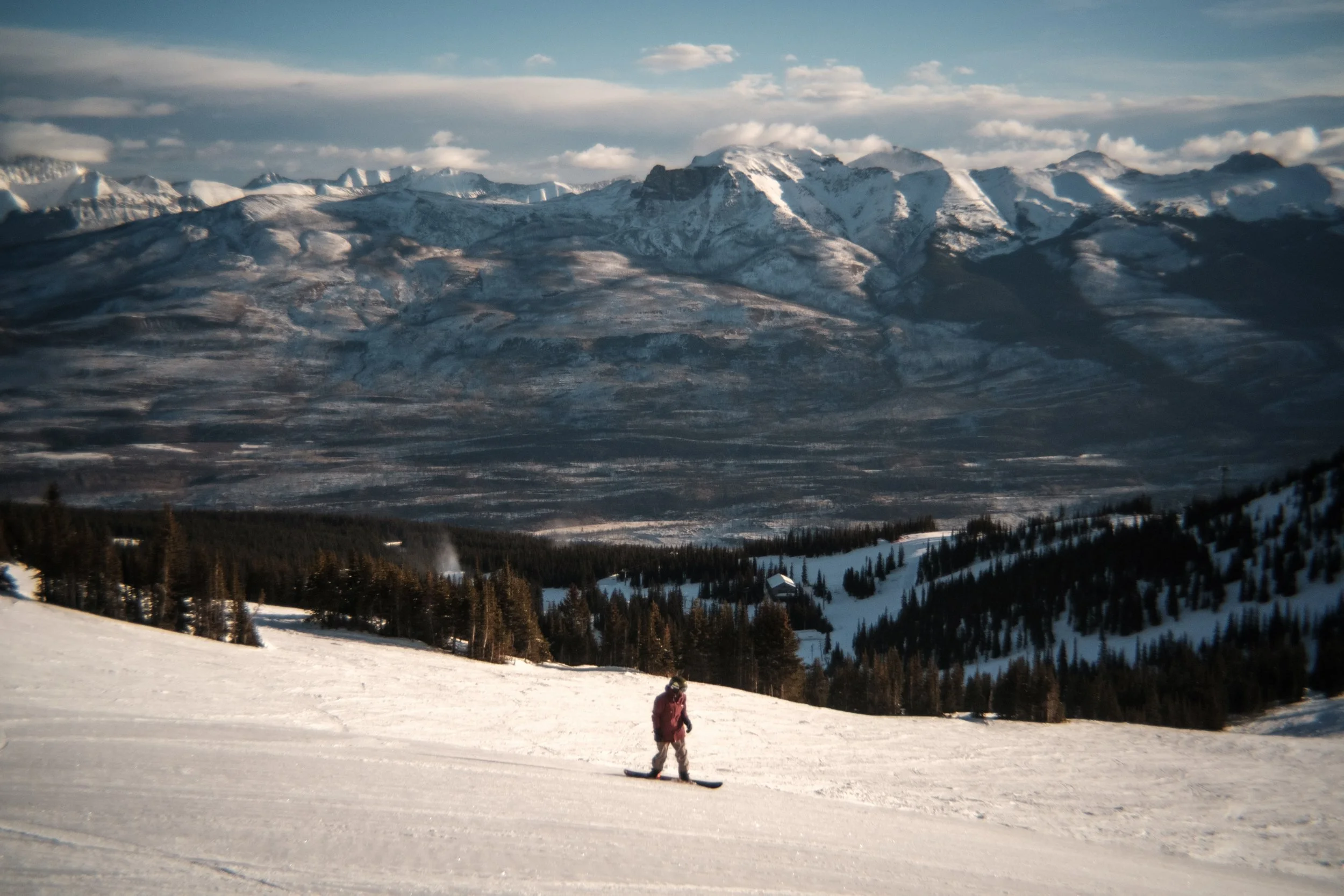 A snowboarder in a red jacket skiing on a snowy slope in a mountainous landscape with snow-covered peaks and evergreen trees.