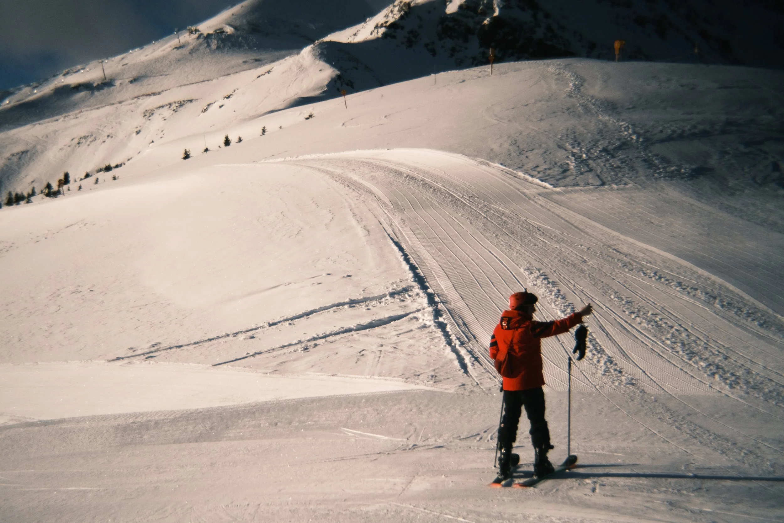 A person in red winter gear and holding ski poles stands on snow, pointing at the ski trail on a mountain slope.
