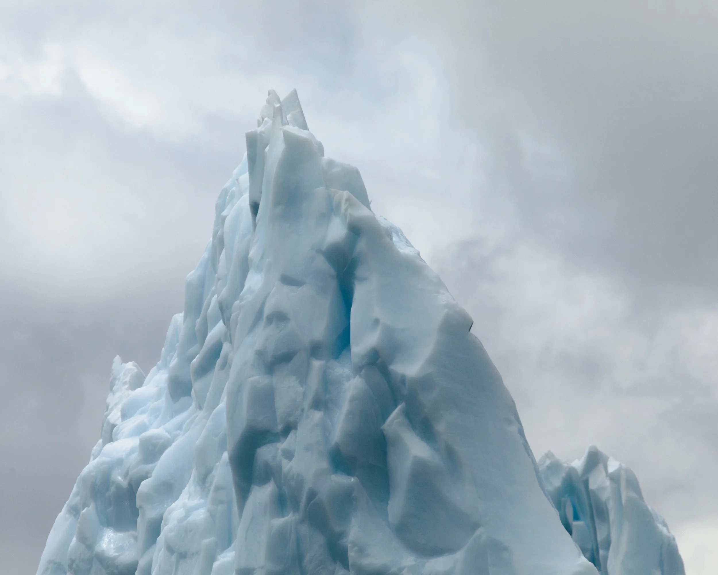 A tall iceberg or glacier ice formation against a cloudy sky.