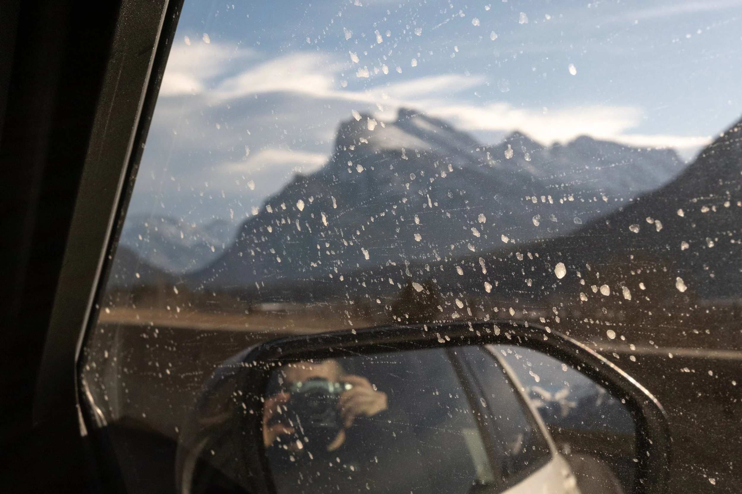 View of snow-capped mountains through a dusty car window and side mirror, with the reflection of the photographer holding a camera.