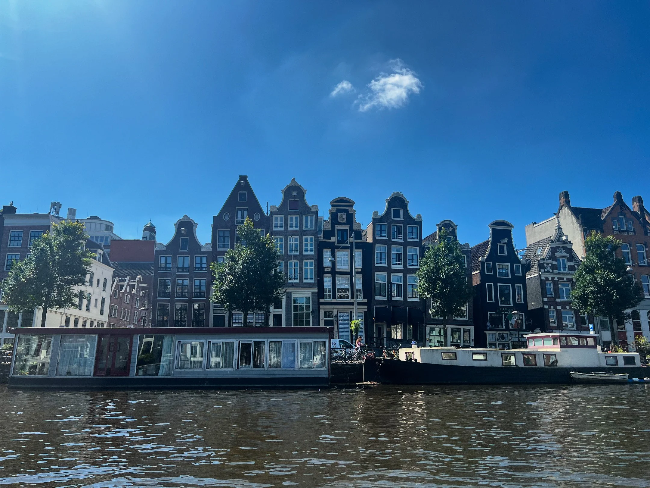 A canal with houseboats in front of a row of traditional Dutch-style buildings and trees under a bright blue sky.