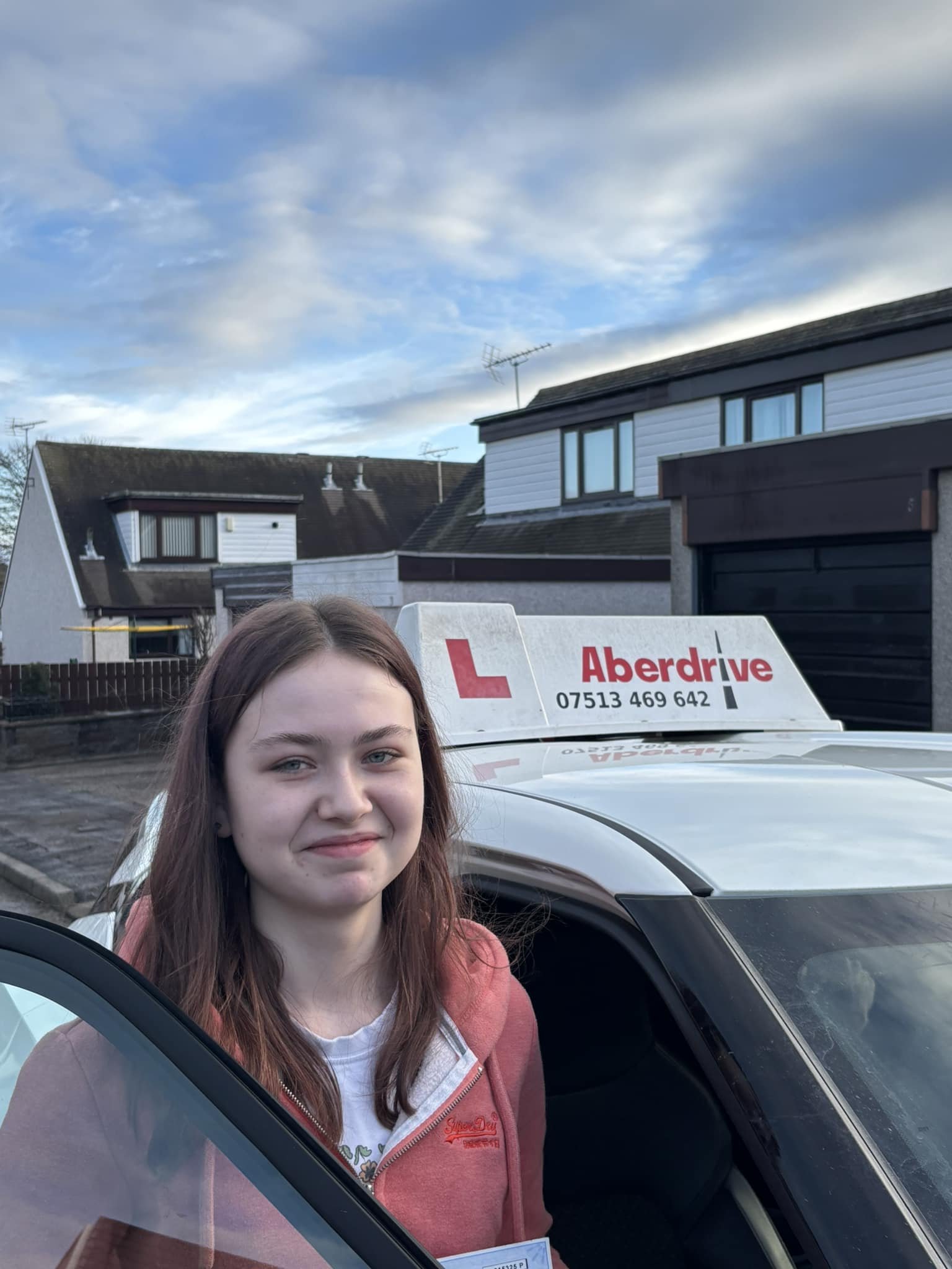 Teen girl with long brown hair standing outside next to driving school car with 'Aberdrive' sign on top, residential neighborhood background, cloudy sky.