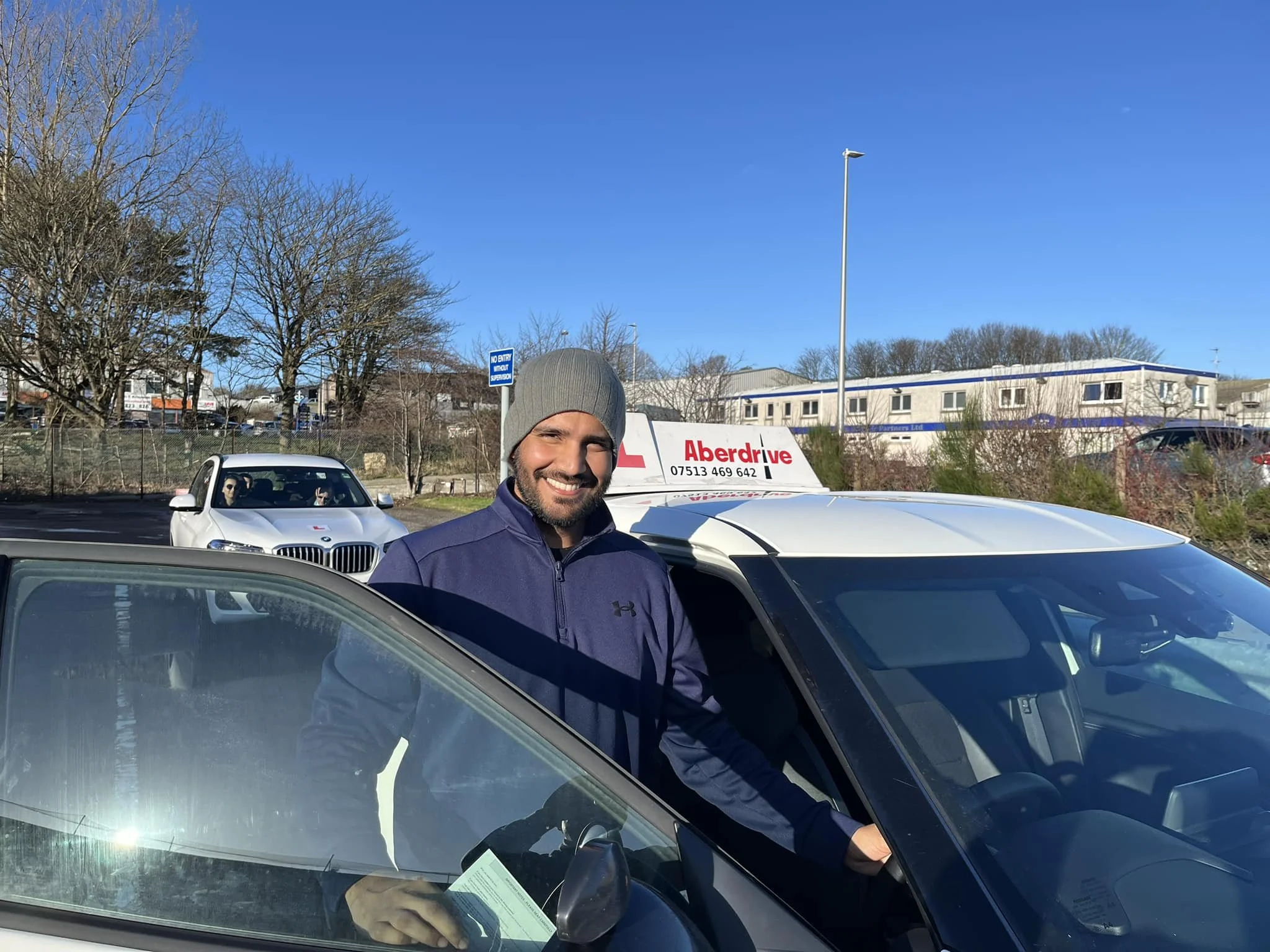 A man in a gray beanie and purple jacket smiling and standing next to the driver's side of a vehicle. In the background, there is a white car with two passengers, some leafless trees, a blue sign, and a white building under a clear blue sky.