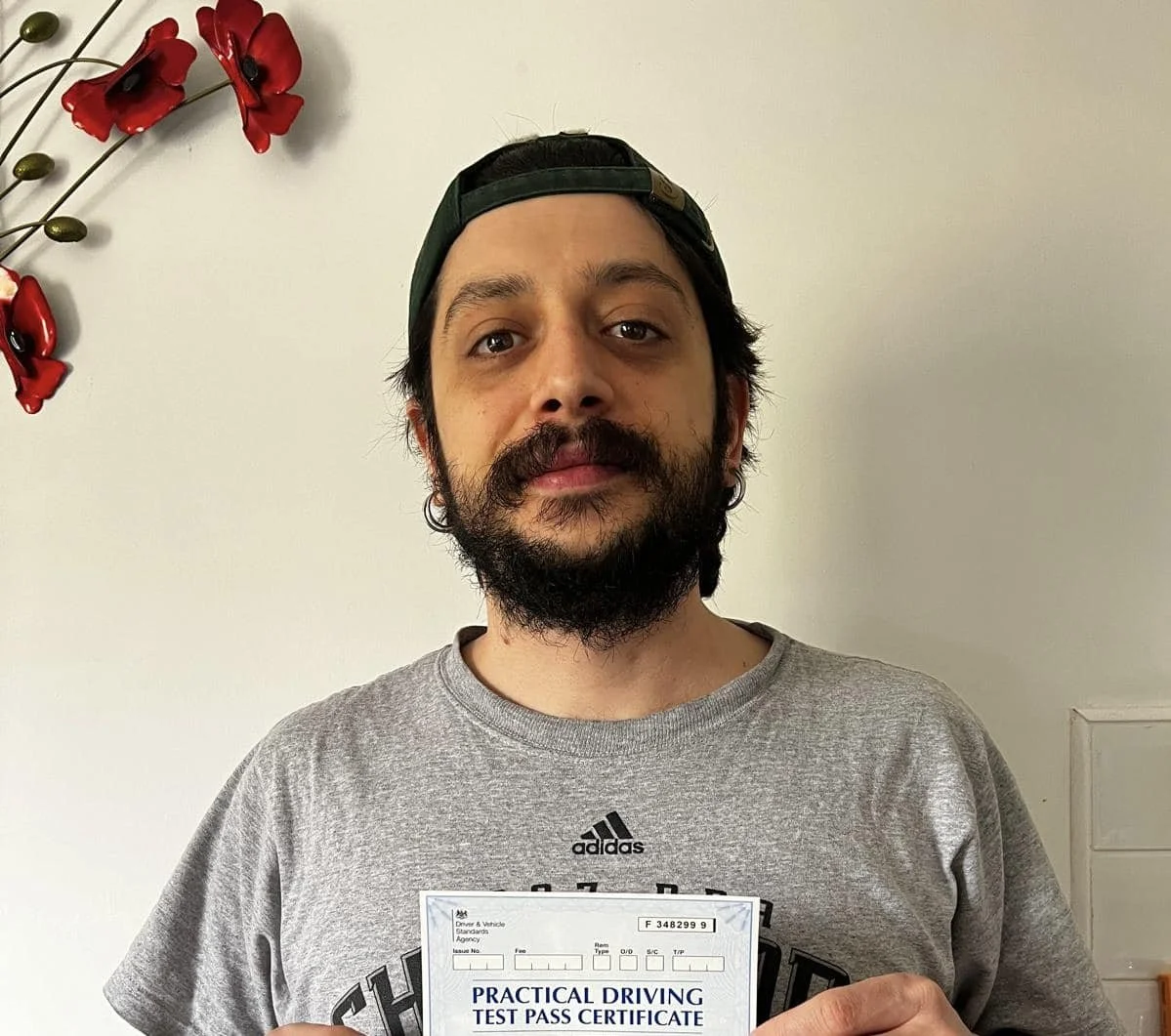 A man with a beard and mustache wearing a gray athletic shirt and a backward dark green cap, holding a practical driving test pass certificate, standing in front of a plain wall with red flower decorations.