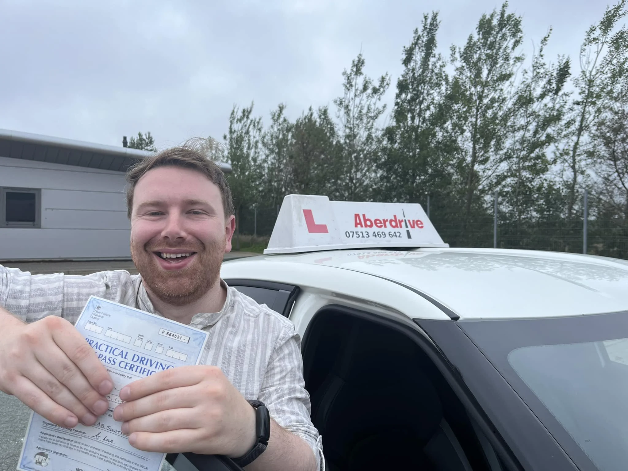 A smiling man holding a practical driving pass certificate standing next to a white driving school car with an 'Aberdrive' sign on top, outdoors with trees and a building in the background.