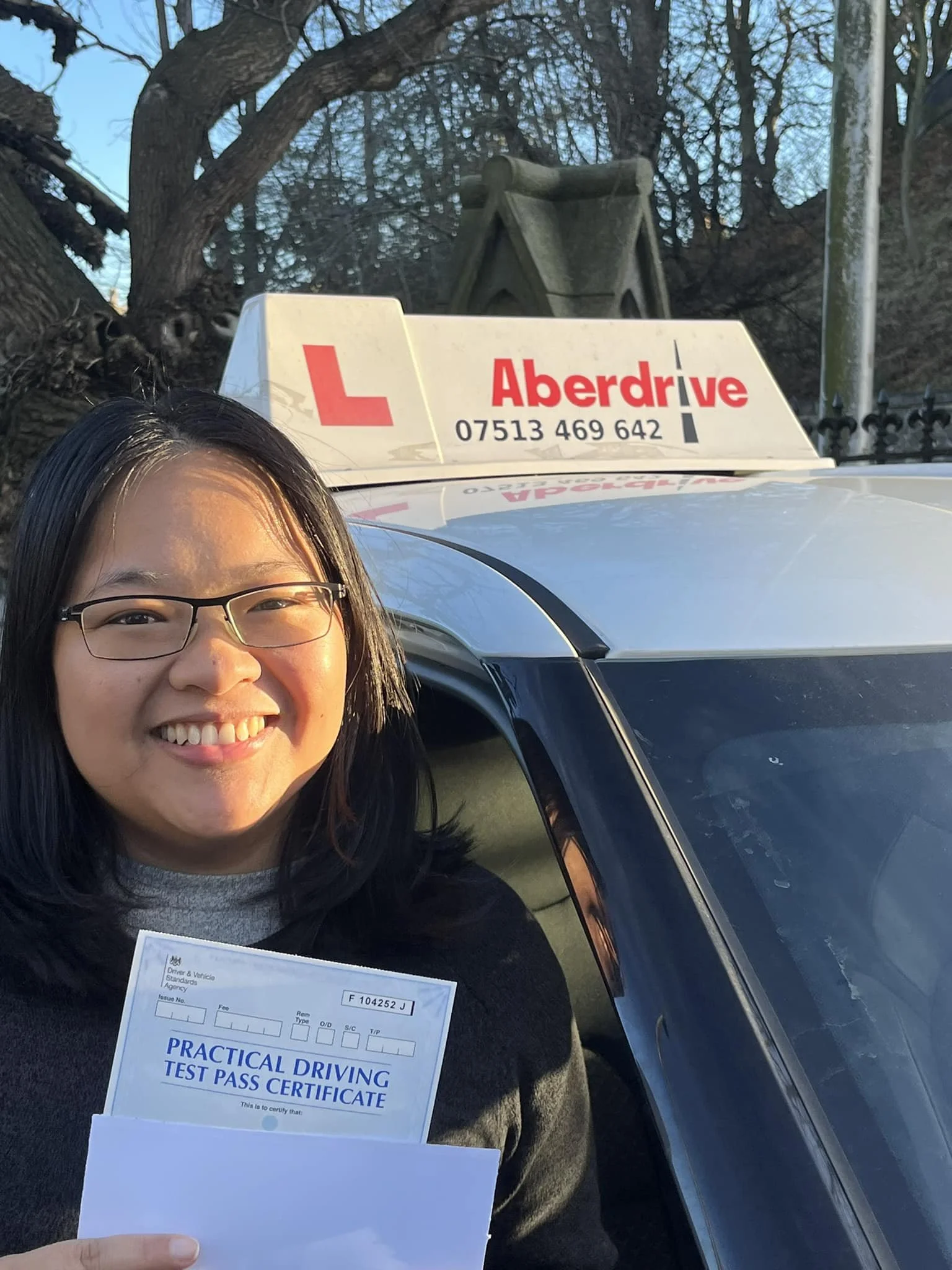 Smiling woman holding a practical driving test pass certificate standing next to a car with an Edinburgh driving school sign on top.