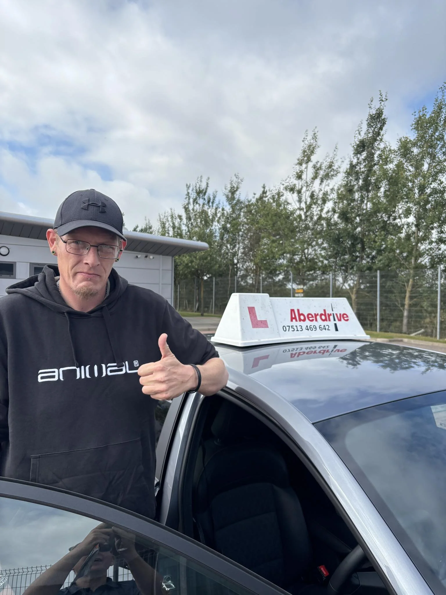 A man in a black hoodie and cap gives a thumbs-up next to a driving school car with an L sign and Aberdeen drive contact information, outdoors under cloudy sky.