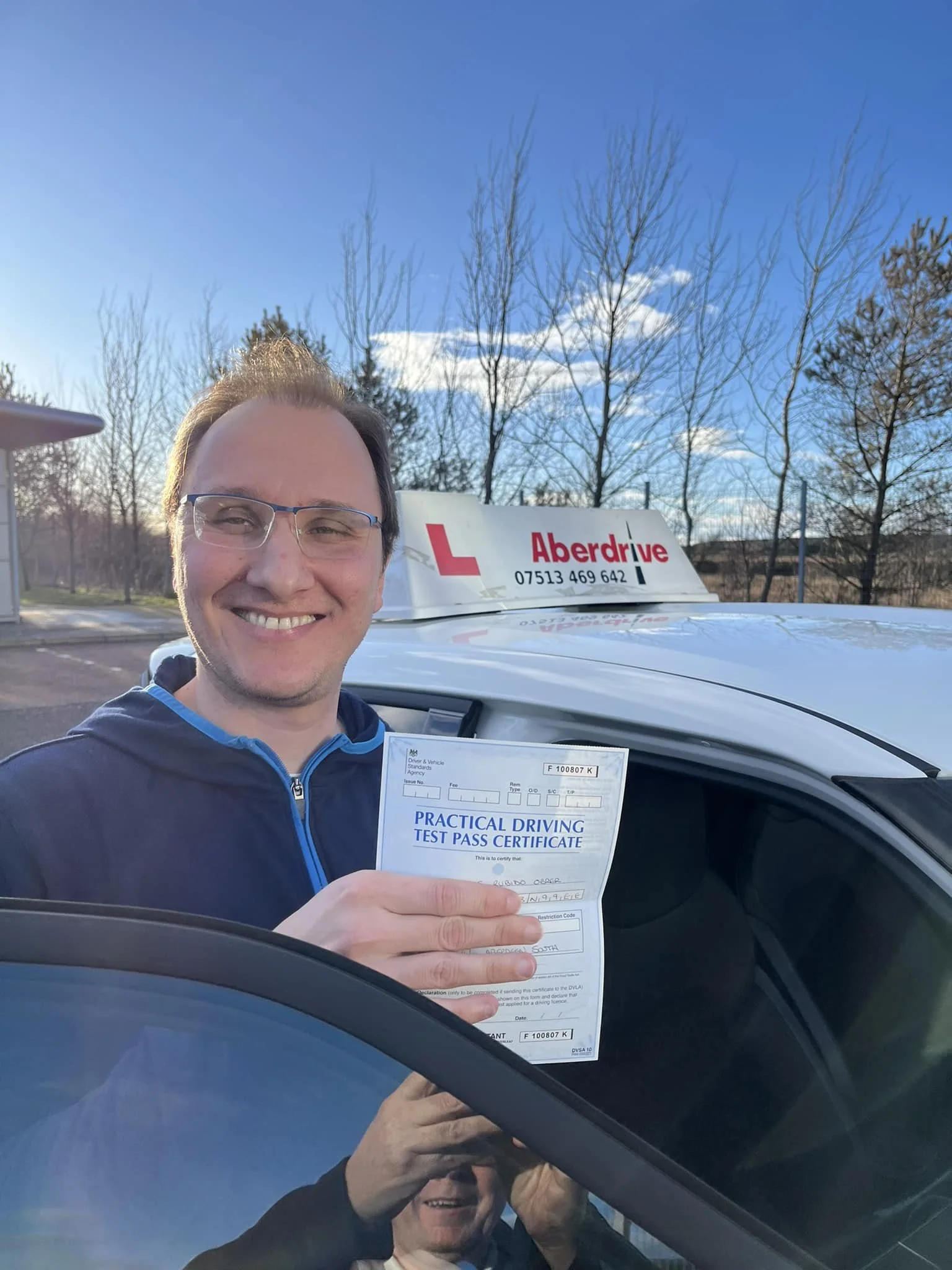 A man smiling and holding a practical driving test pass certificate stands in front of a driving school car with a roof sign reading "Aberdrive." The background shows a clear sky and leafless trees.