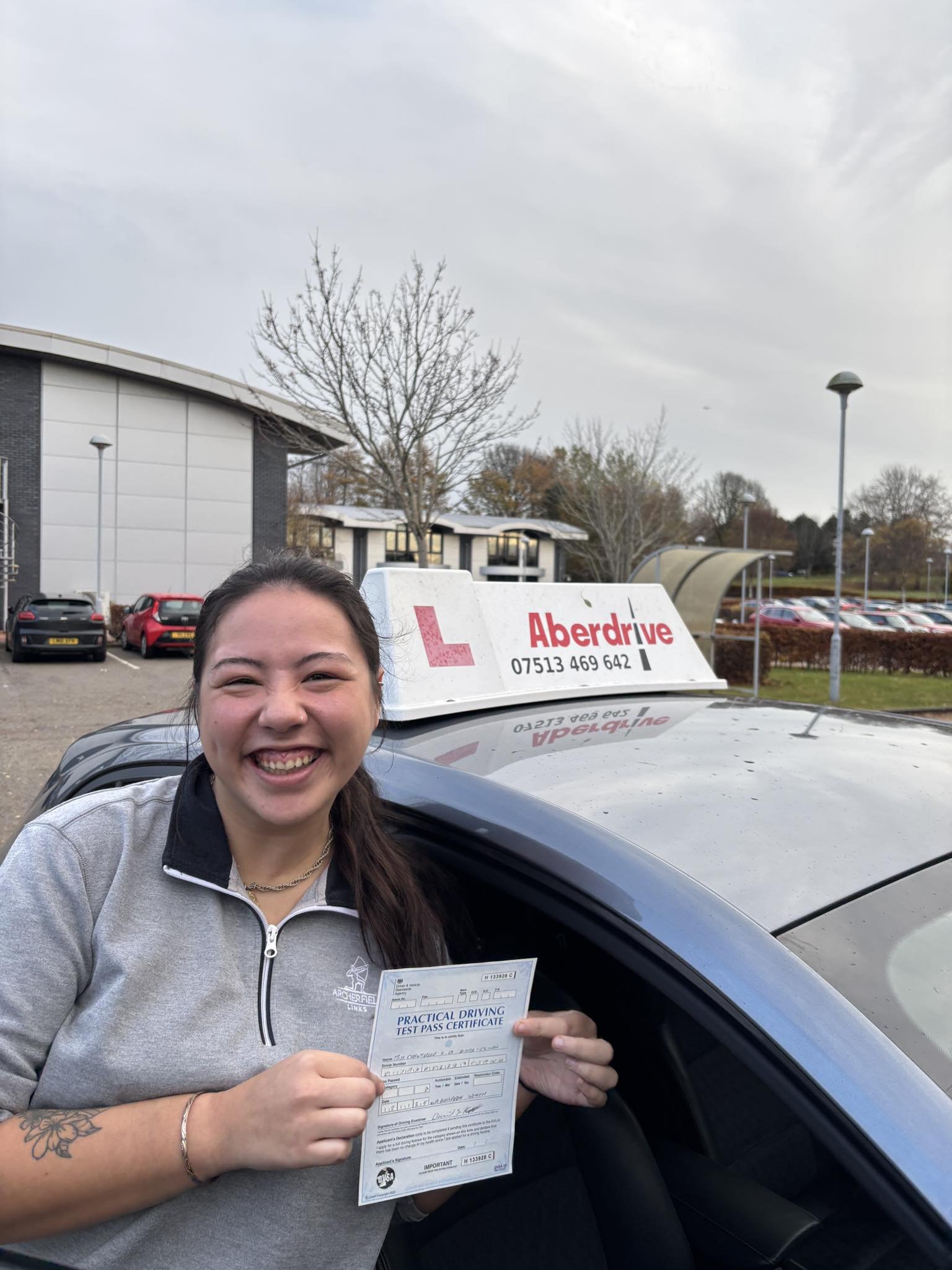 A smiling woman holding a practical driving test pass certificate inside a car, with a driving school sign on the roof and a parking lot in the background.