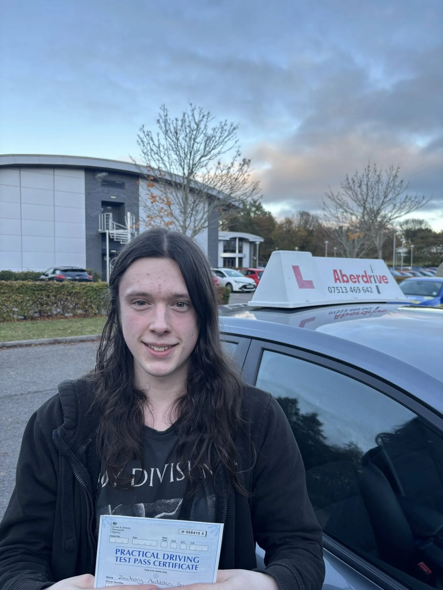 A young person outdoors holding a practical driving test pass certificate in front of a car with a driving school sign on top that reads 'Aberdrive'.