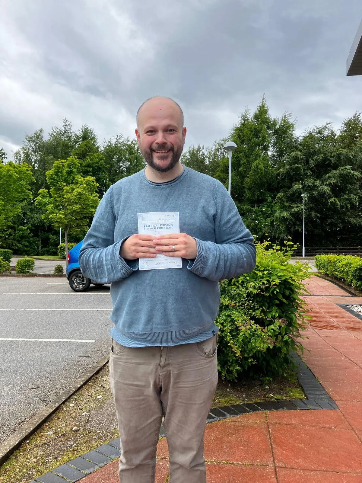 A man standing outside in front of greenery, holding a driver's license and smiling.