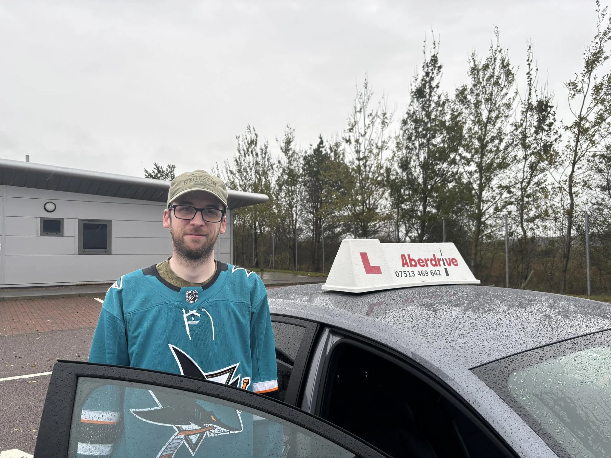 A man standing next to a driving school car, wearing glasses and a hockey jersey, with a canopy and trees in the background on a cloudy day.