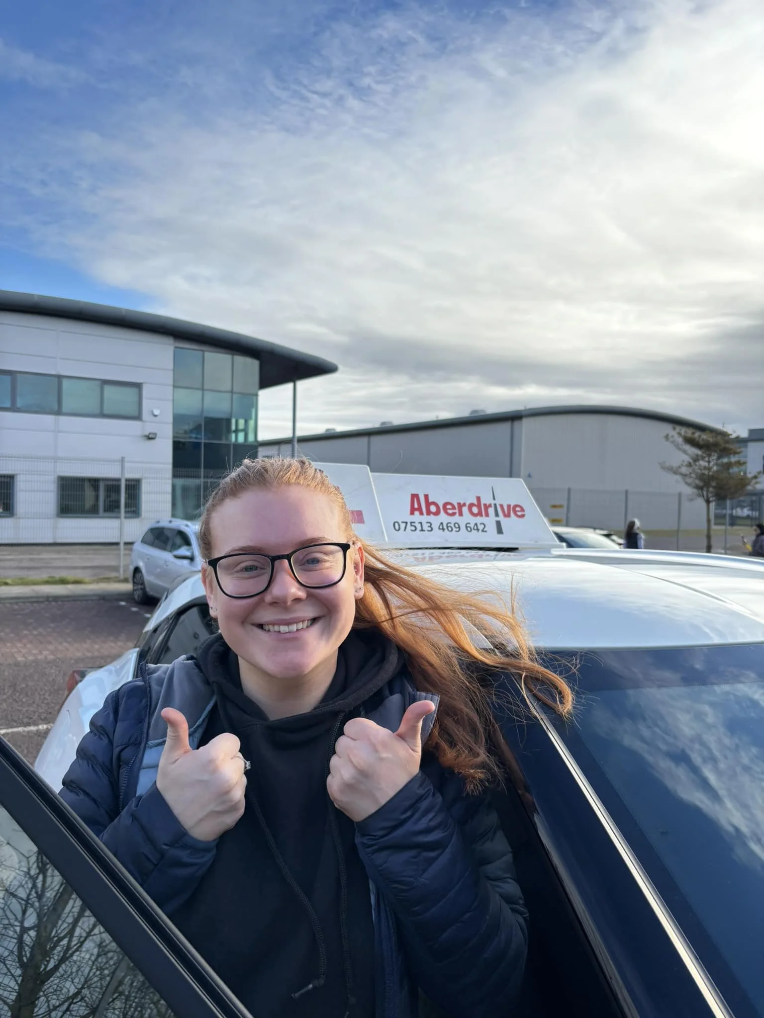 A smiling woman with glasses and red hair giving a thumbs-up outside in front of a white vehicle with a sign that reads 'Aberdrive' and a phone number. The background includes a modern building and a partly cloudy sky.