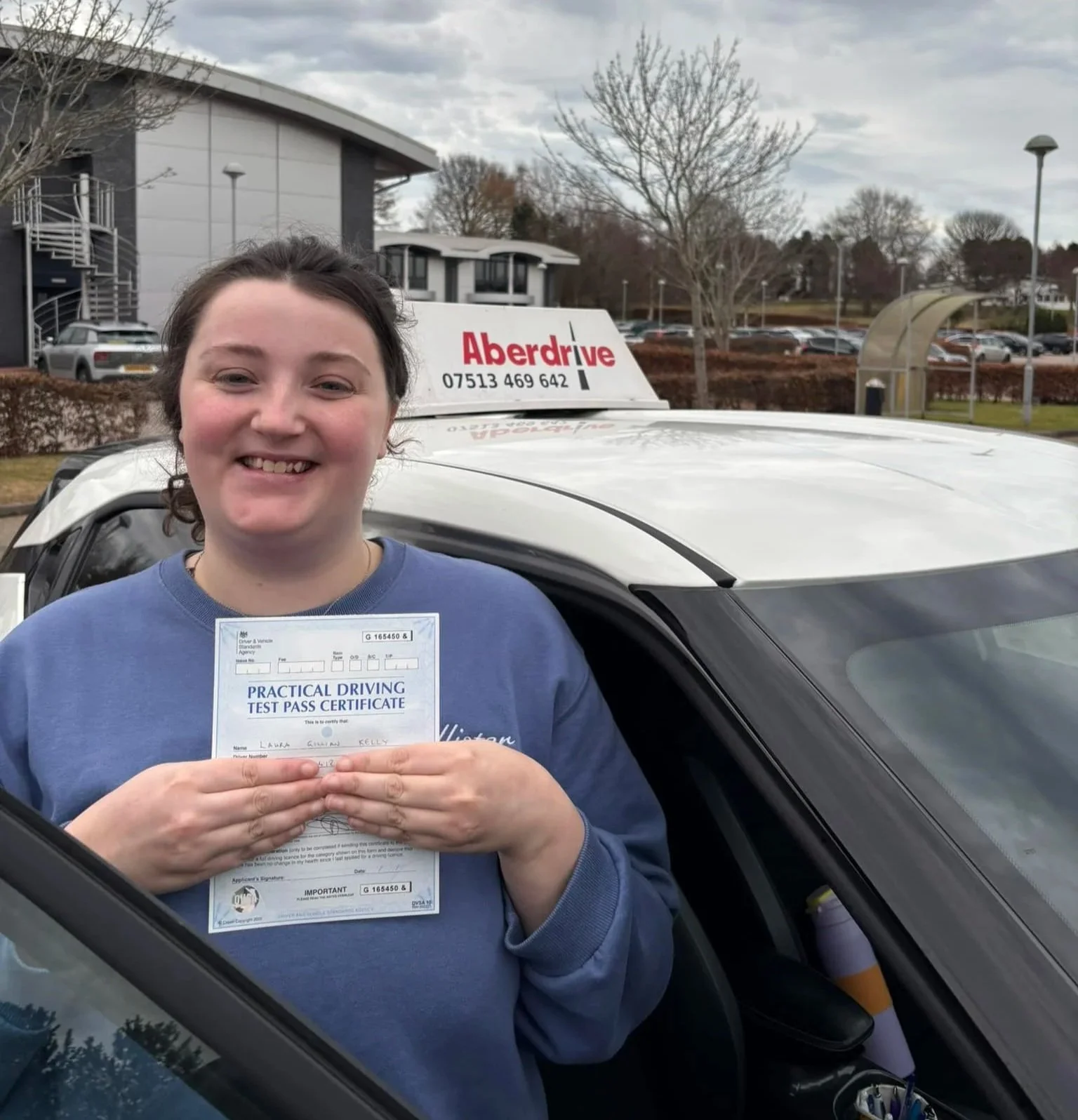 A woman smiling and holding a practical driving test pass certificate while sitting in a car. The car is parked outdoors with trees and a building in the background, and a sign on the car roof reads 'Aberdrive' with a phone number.