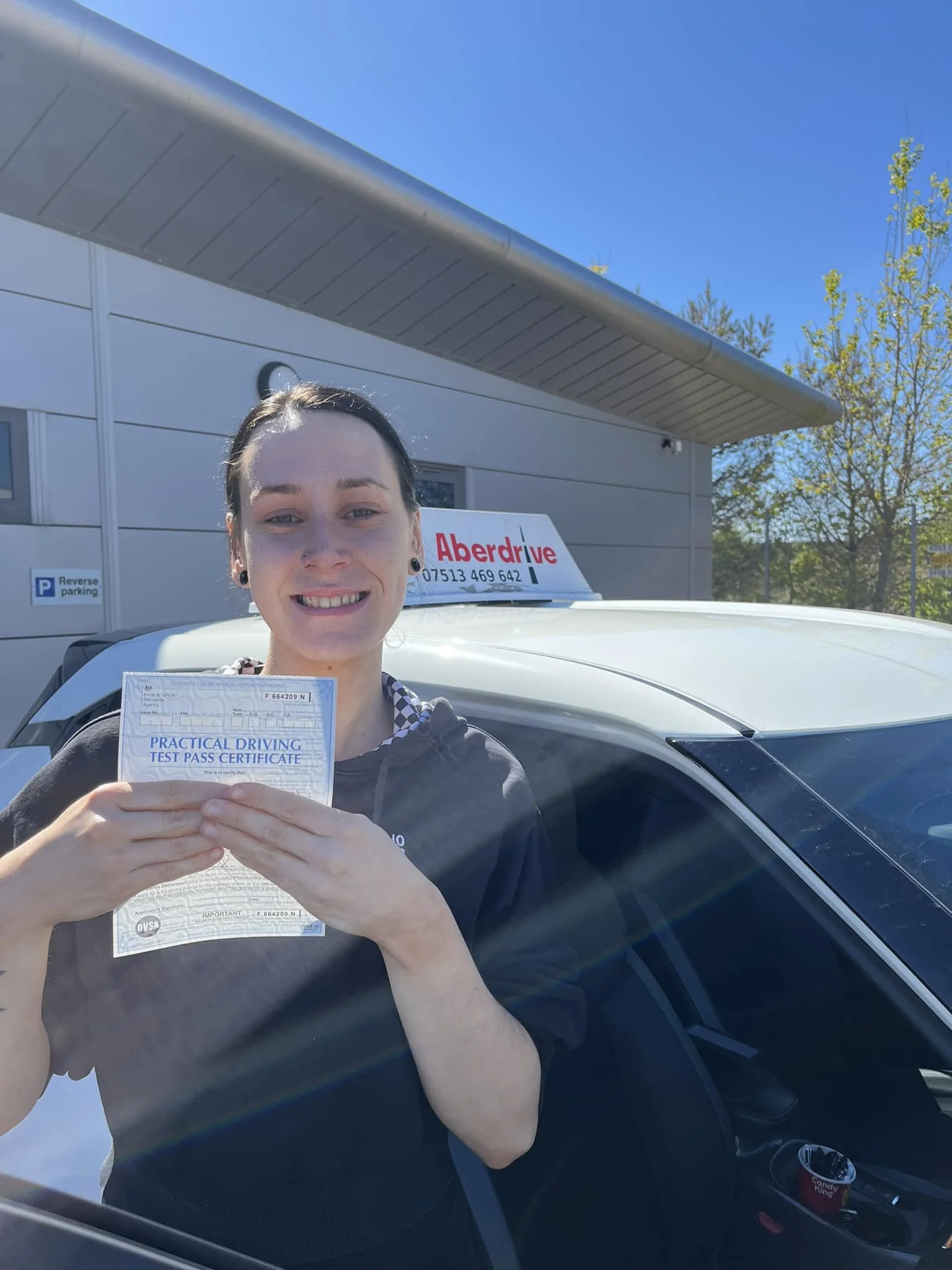Young woman with dark hair and earrings holding a practical driving test pass certificate, smiling in front of a car with an Aberdrive sign, outside a building with trees and clear blue sky.