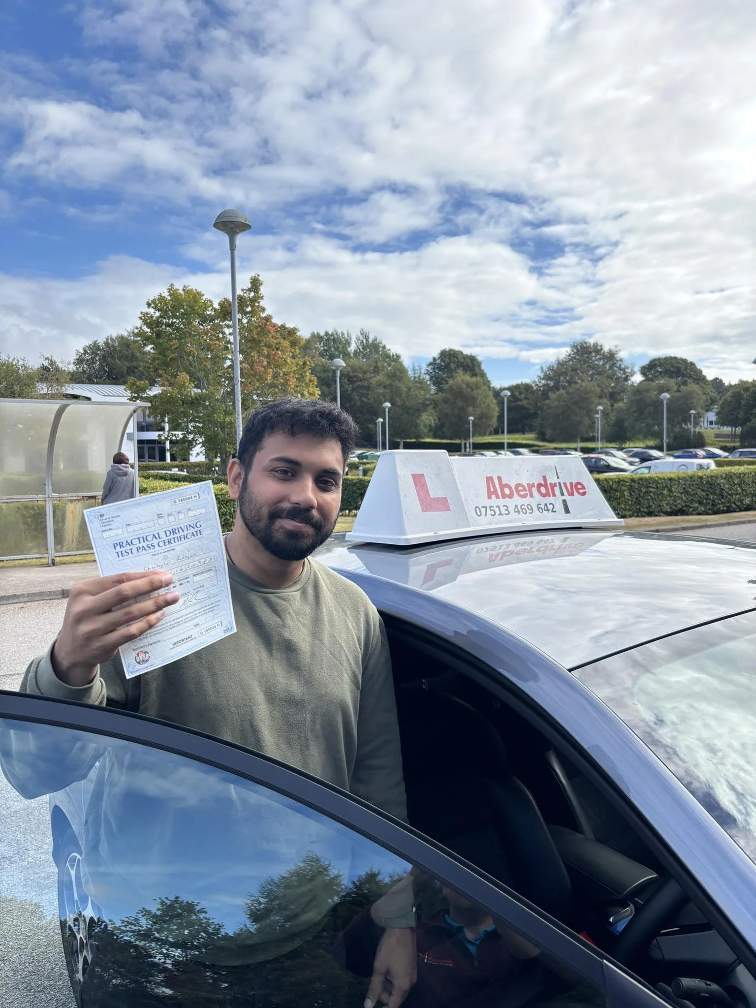 A man holding a practical driving test pass certificate next to a driving school car with an 'Aberdrive' sign on top, in a parking lot with trees and cars in the background.