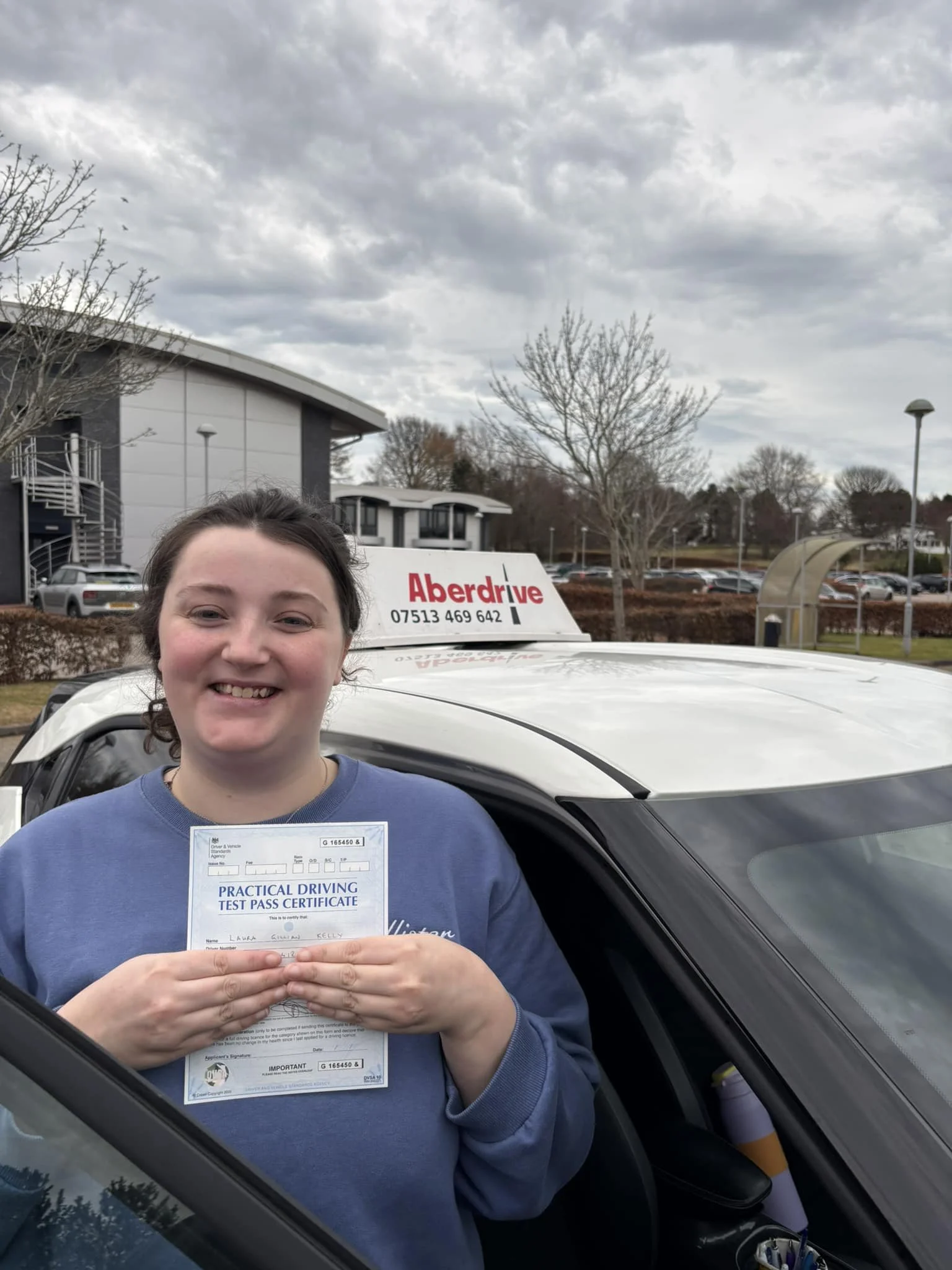 A young woman holding a practical driving test pass certificate, smiling, standing next to a white car with an 'Aberdrive' sign on the roof in a parking lot on an overcast day.